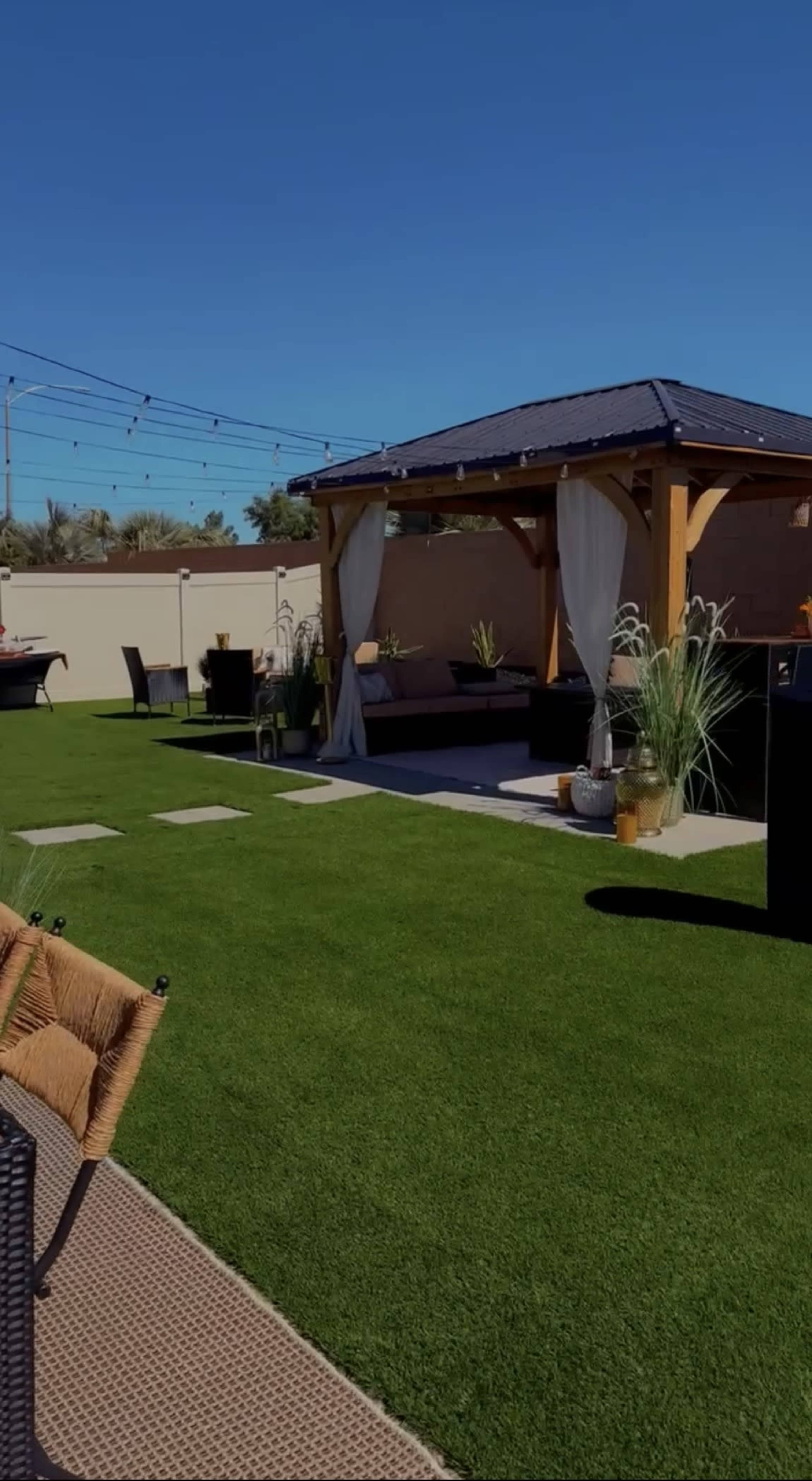 The image shows a landscaped backyard with artificial grass, a wooden gazebo, patio furniture, and a barbecue grill.