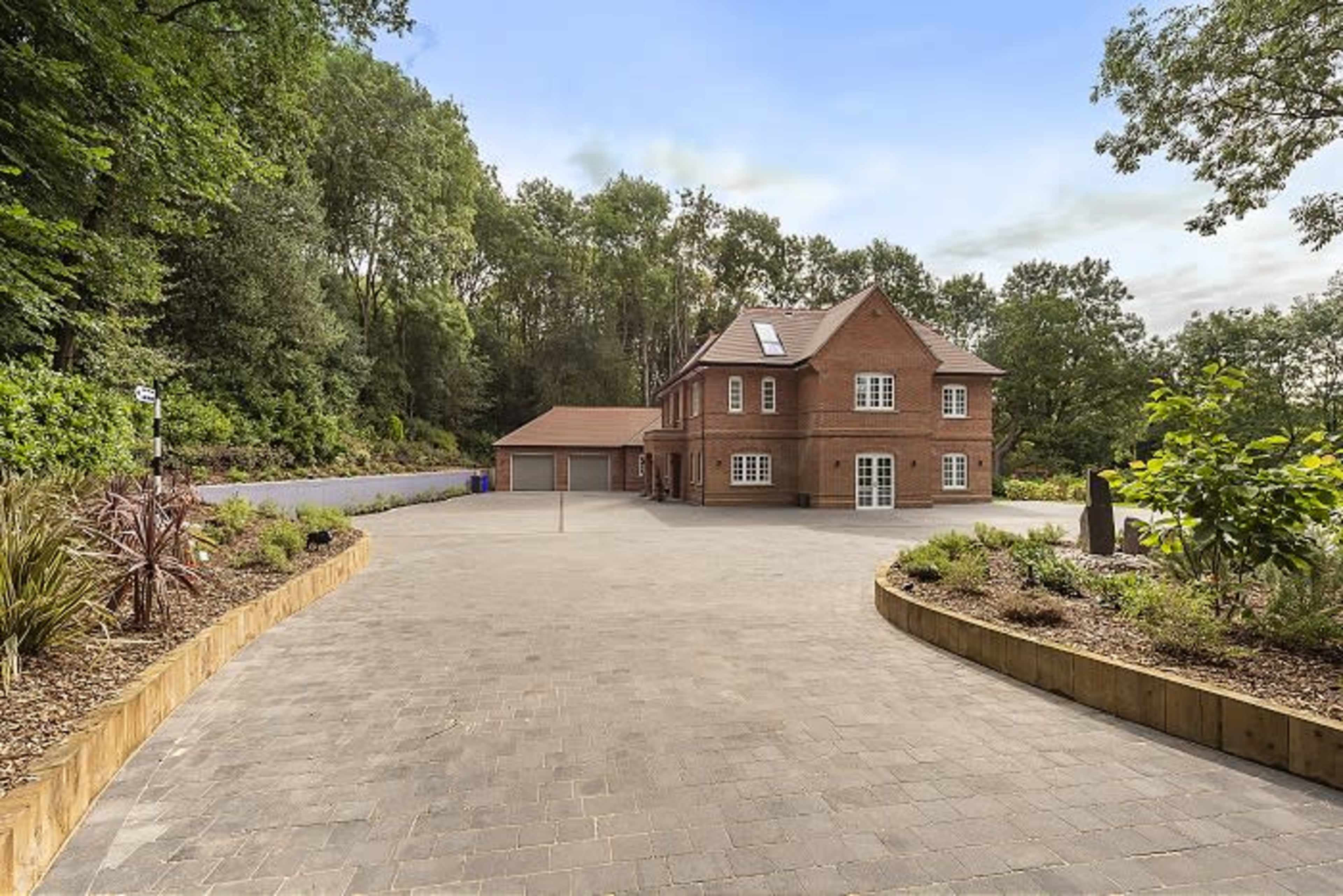 A modern brick house with a large driveway is surrounded by greenery and trees.