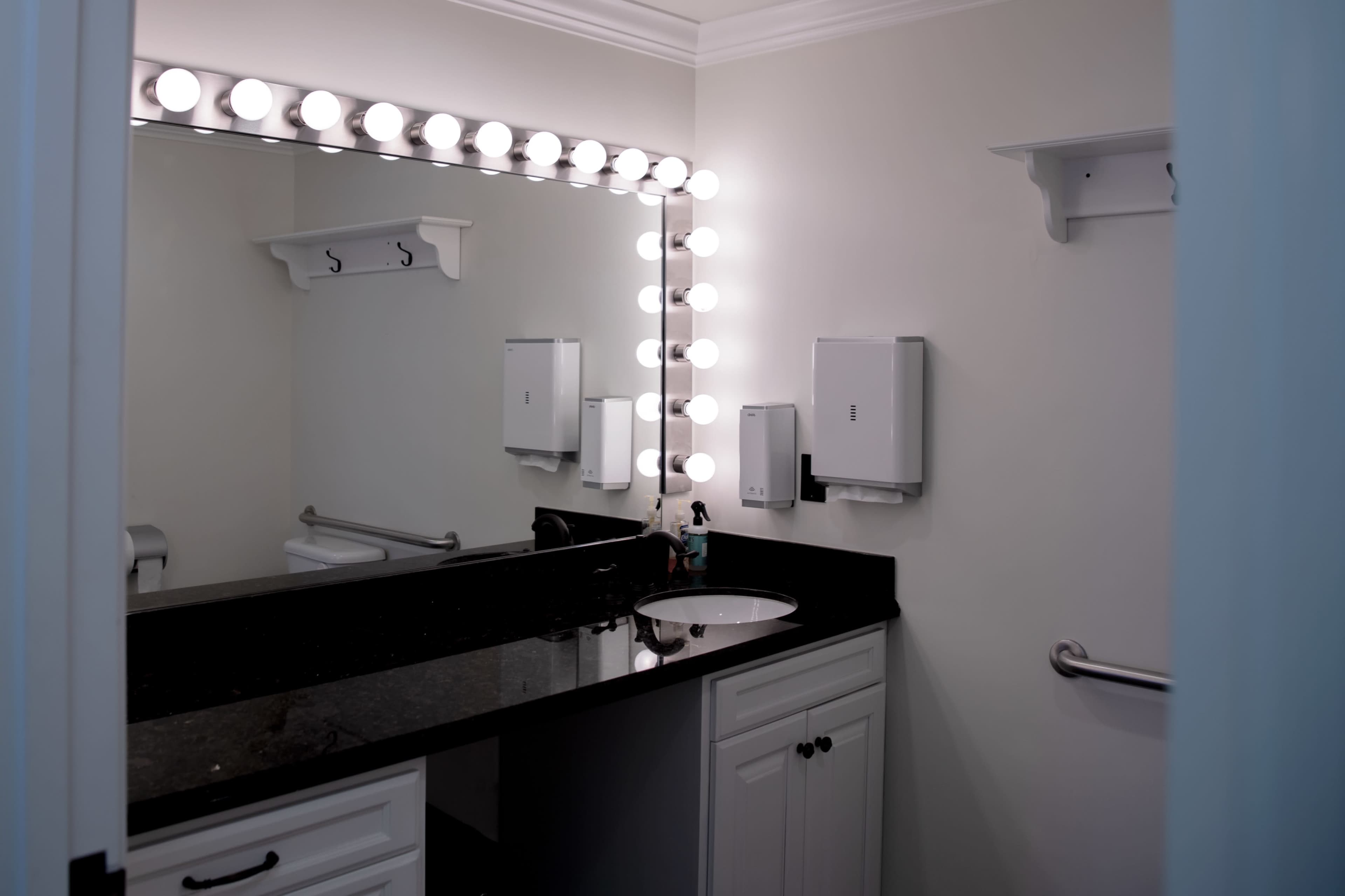 The image shows a well-lit bathroom with a large mirror, black countertop, and white cabinetry.