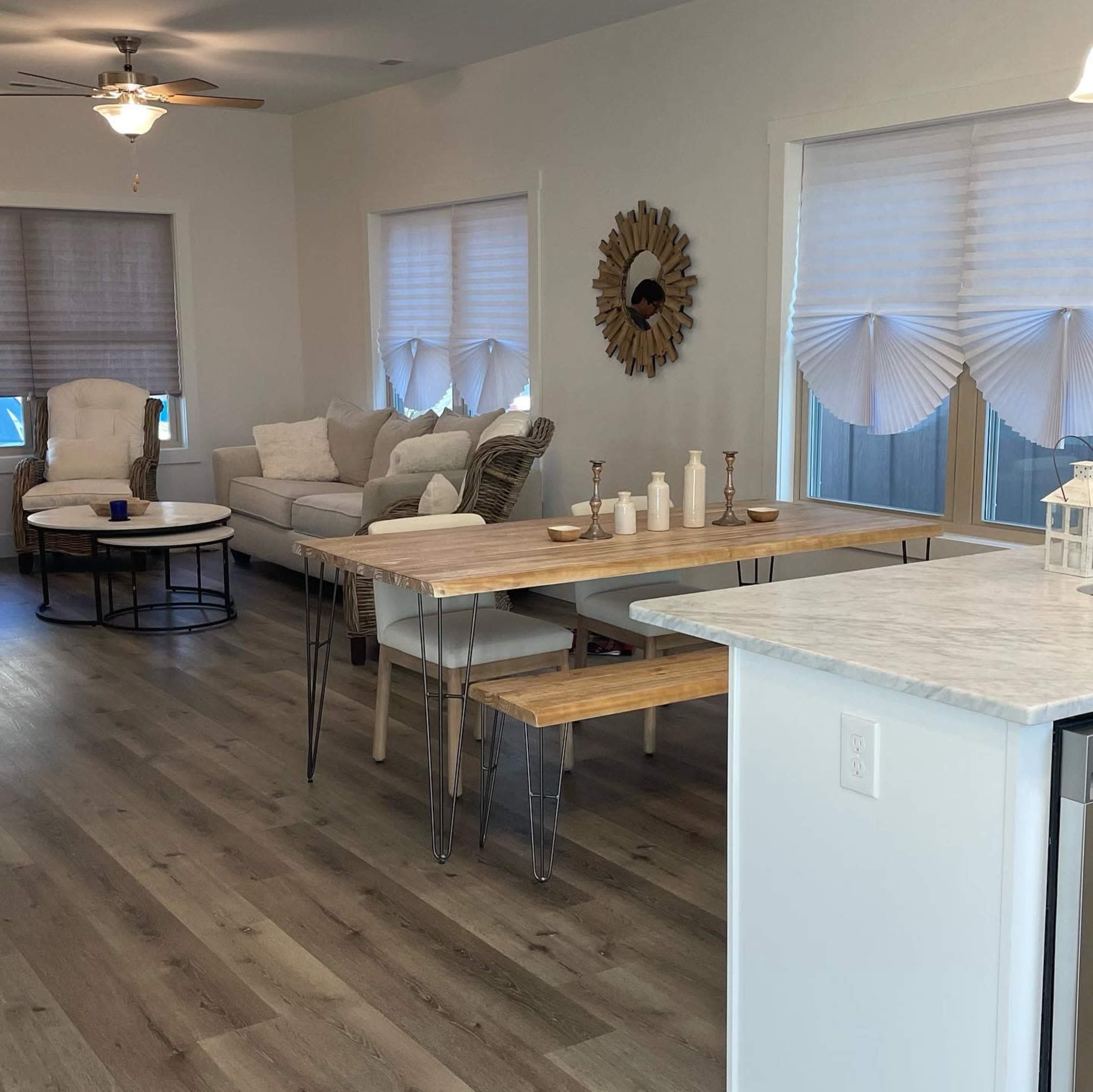 The image shows a modern living space featuring a light-colored sofa set, a circular coffee table, a wooden dining table with metal legs, and large windows with light-colored shades.