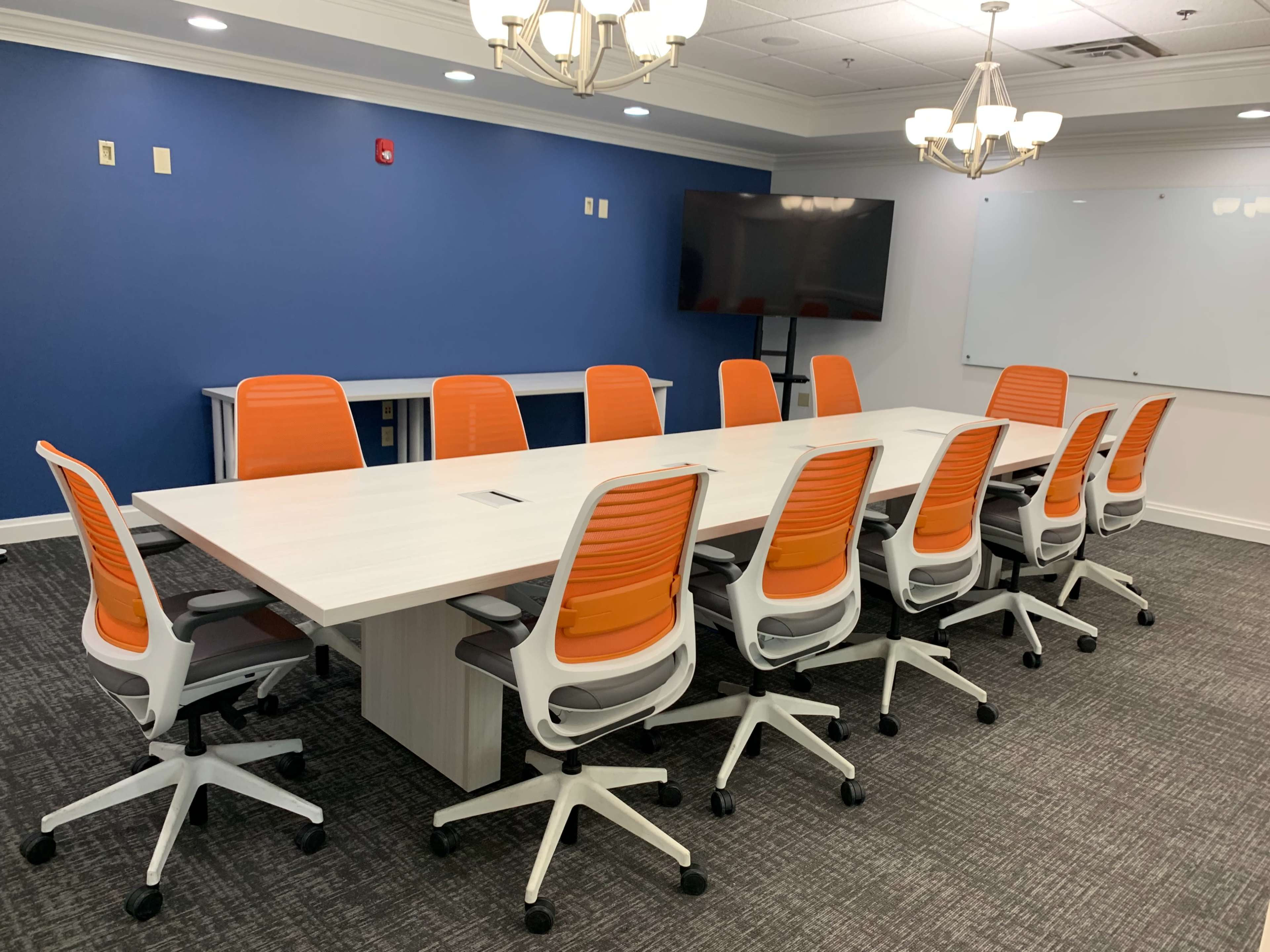 A conference room features a long table surrounded by ten orange and white chairs, with a TV and whiteboard against a blue wall.