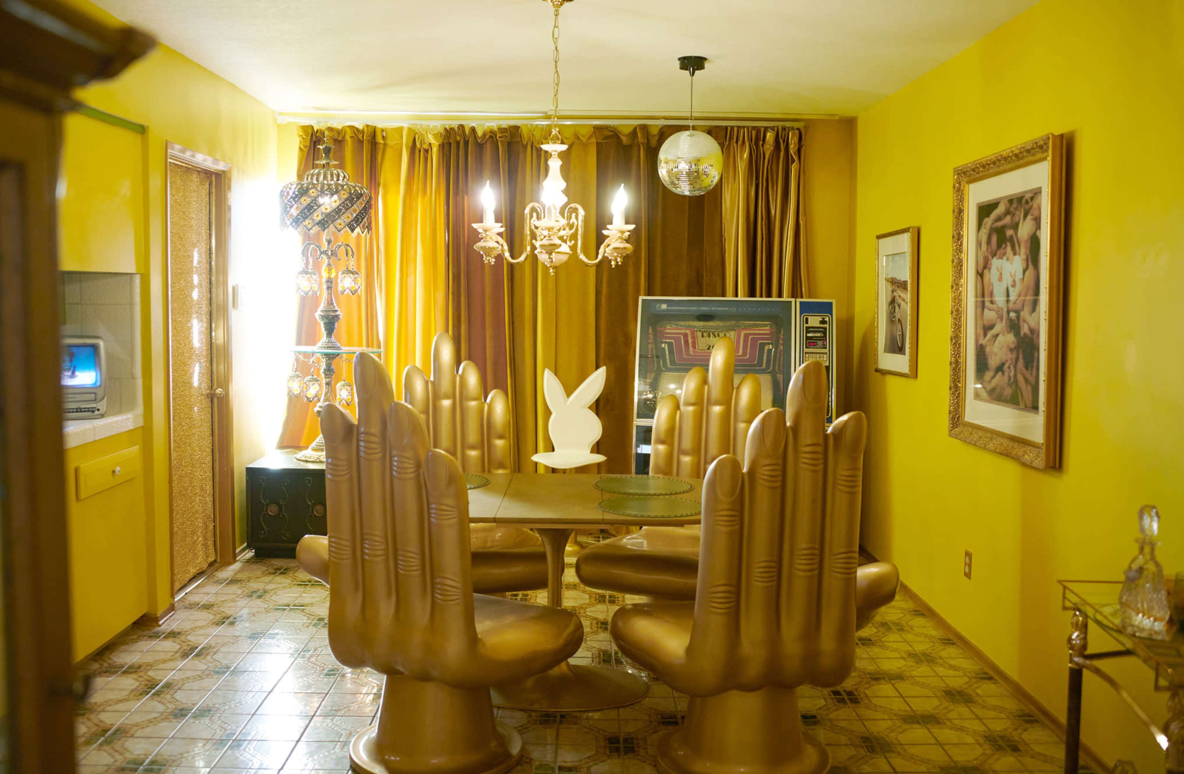 A yellow-colored dining room featuring a table surrounded by large, gold hand-shaped chairs, with a chandelier and a disco ball overhead.