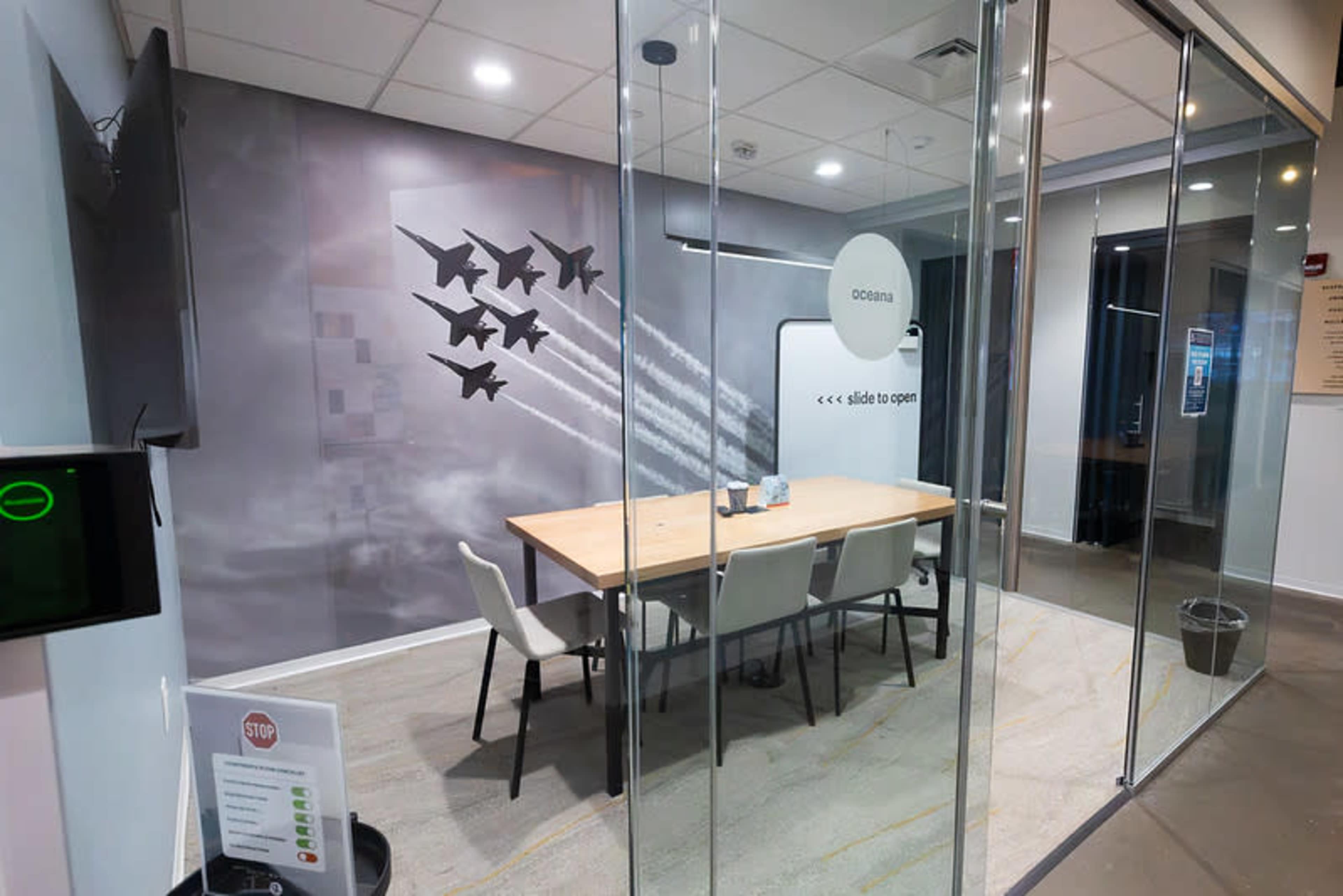 The image shows a modern conference room with a long wooden table surrounded by white chairs, glass walls, and a mural featuring jets flying in formation.