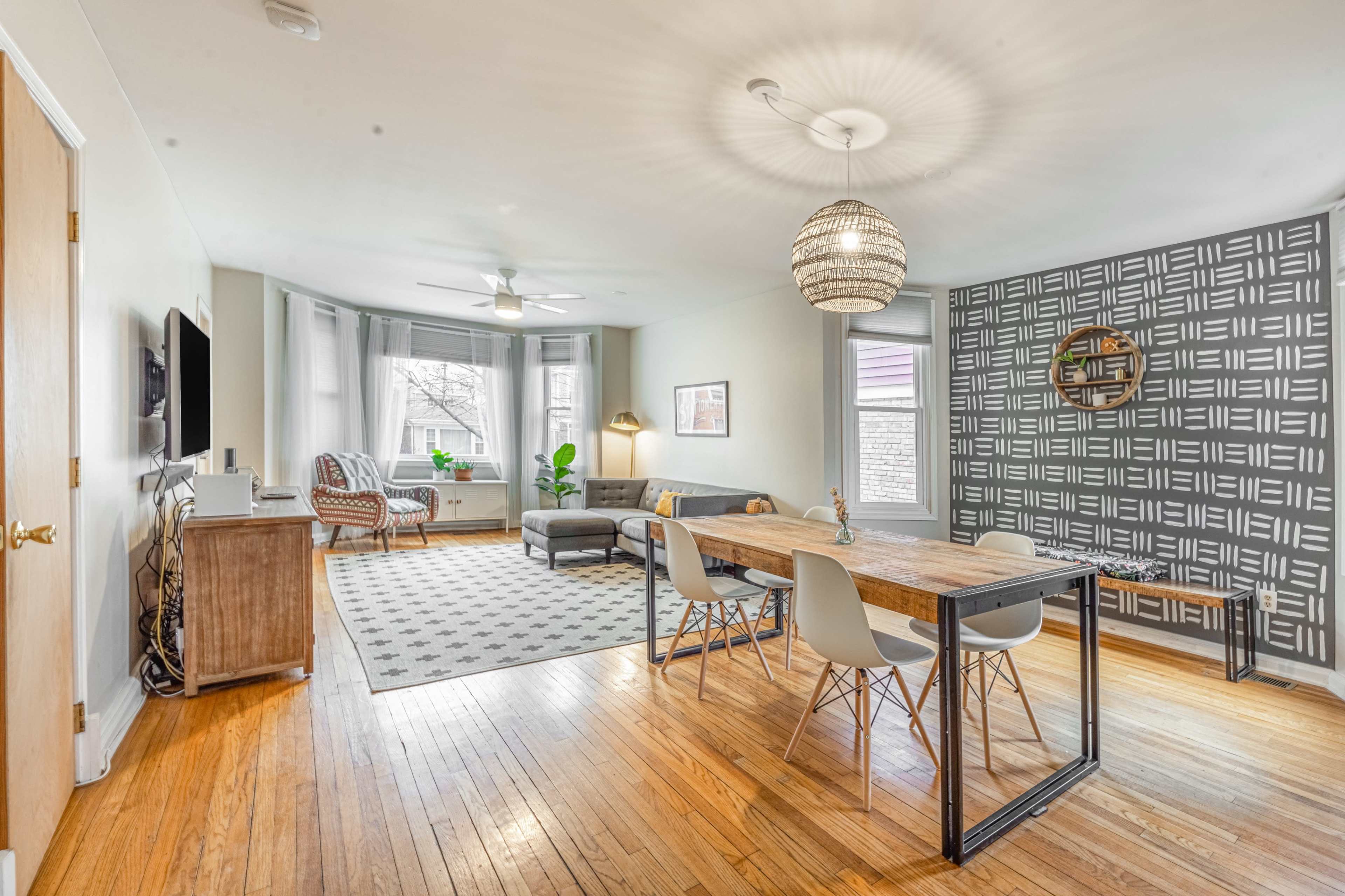 A spacious living room and dining area featuring a wooden table, gray chairs, a patterned rug, and large windows with natural light.