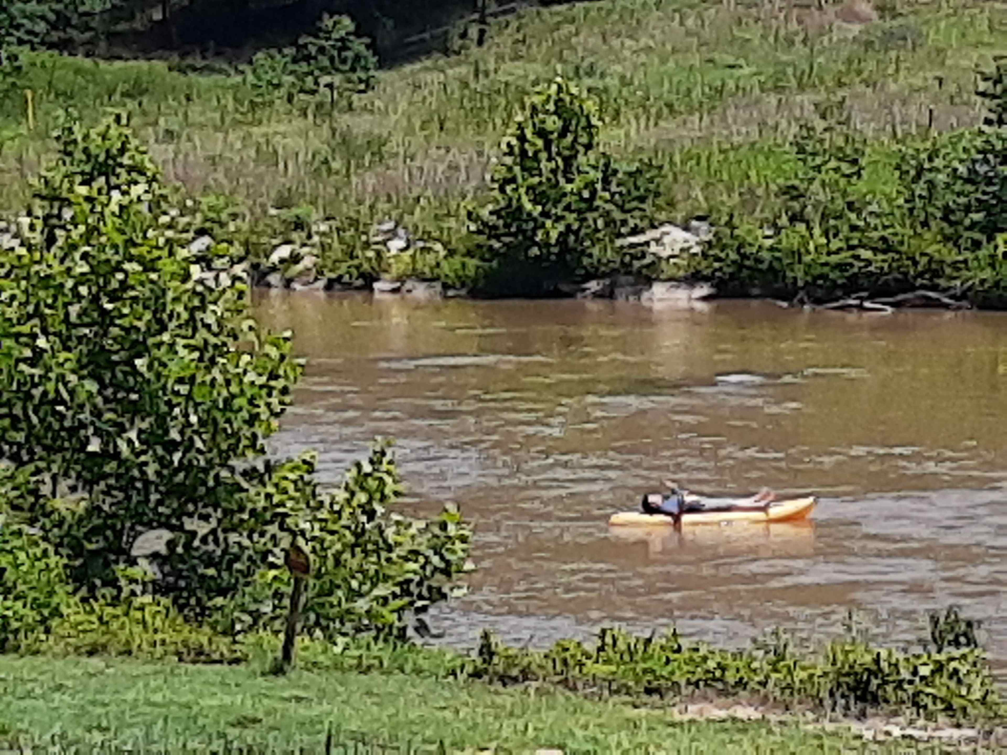 A person lies on a kayak in a brown river surrounded by greenery and rocks.