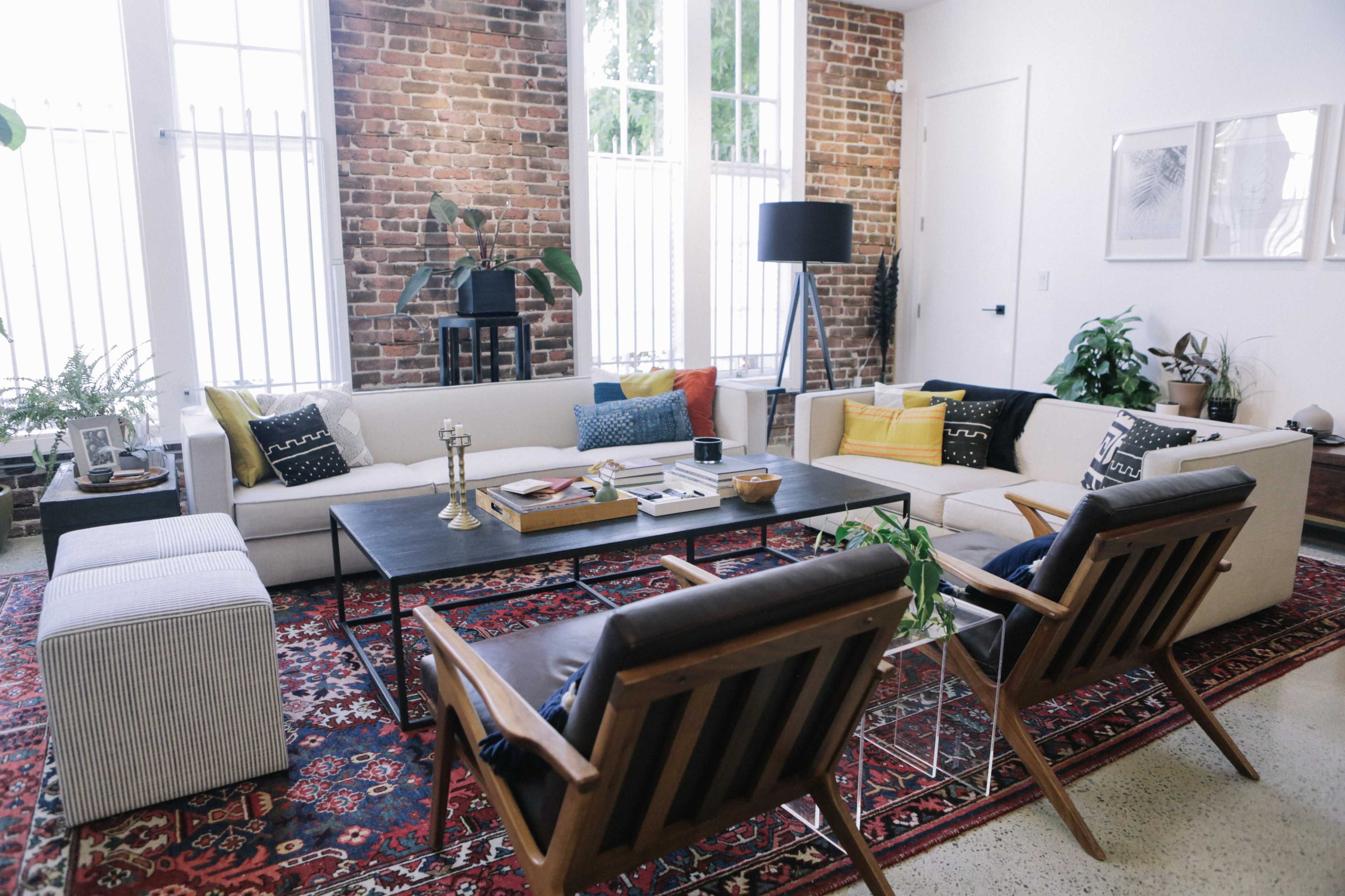 A modern living room features a mix of seating options, including a sectional sofa and two wooden chairs, arranged around a central coffee table on a patterned rug against a backdrop of exposed brick walls and large windows.