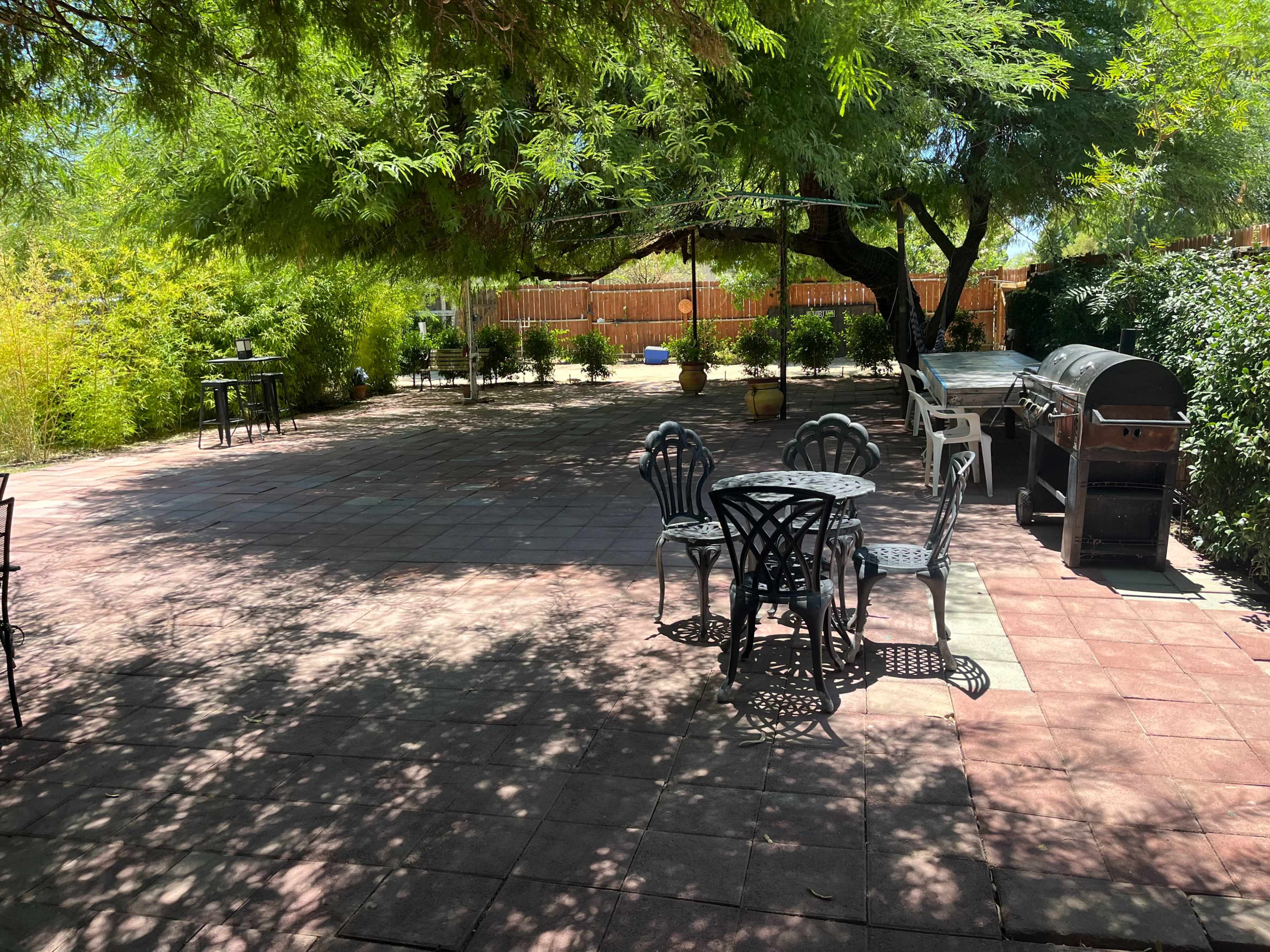 The image shows a spacious outdoor patio area with tiled flooring, shaded by trees, and equipped with metal furniture and a barbecue grill.