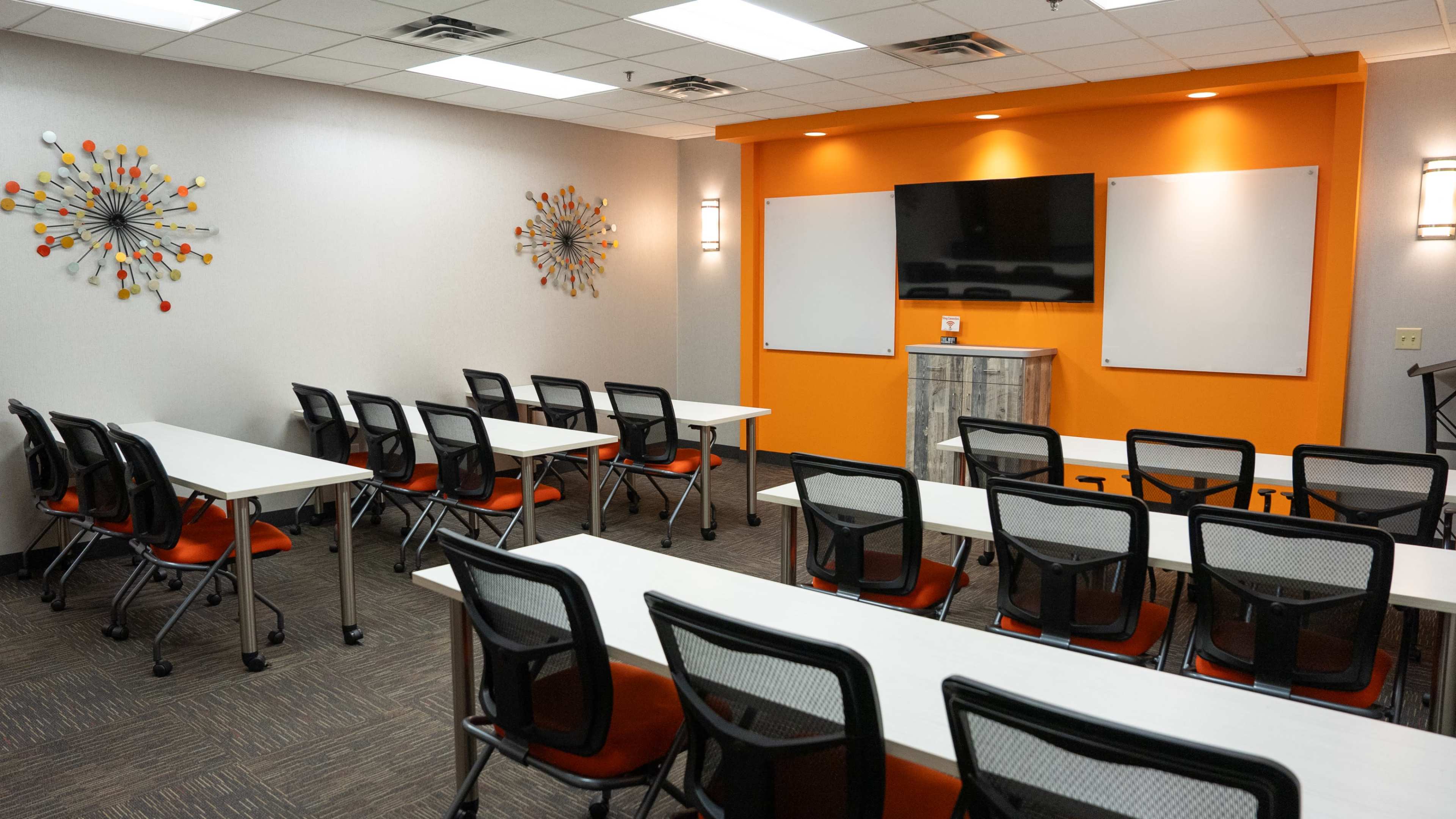 The image shows a well-lit conference room featuring multiple rows of tables and chairs, with walls decorated in orange and gray, and a large screen mounted at the front.