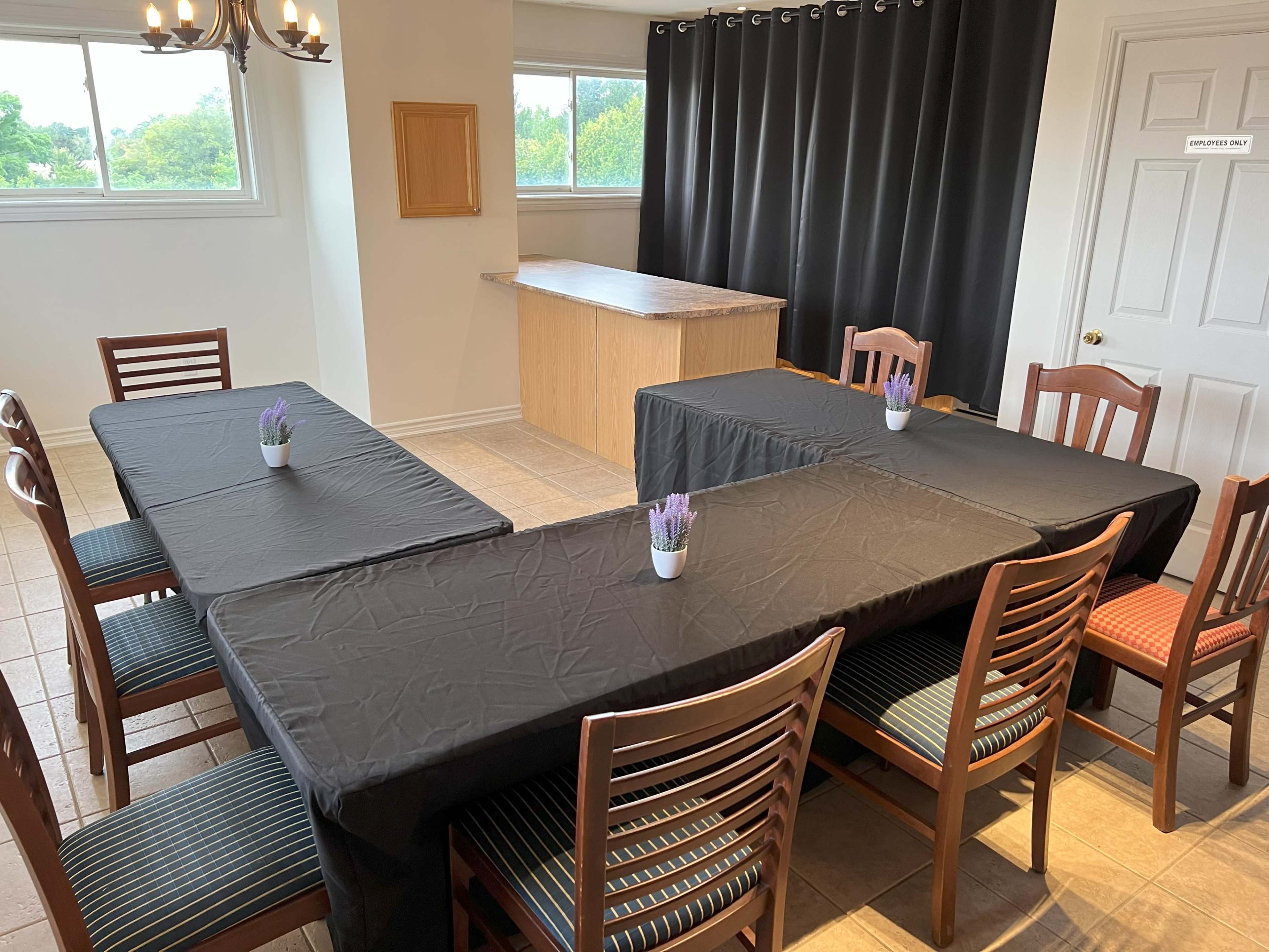 A room with four black-covered tables arranged in a rectangular layout, surrounded by wooden chairs and featuring a countertop in the background.