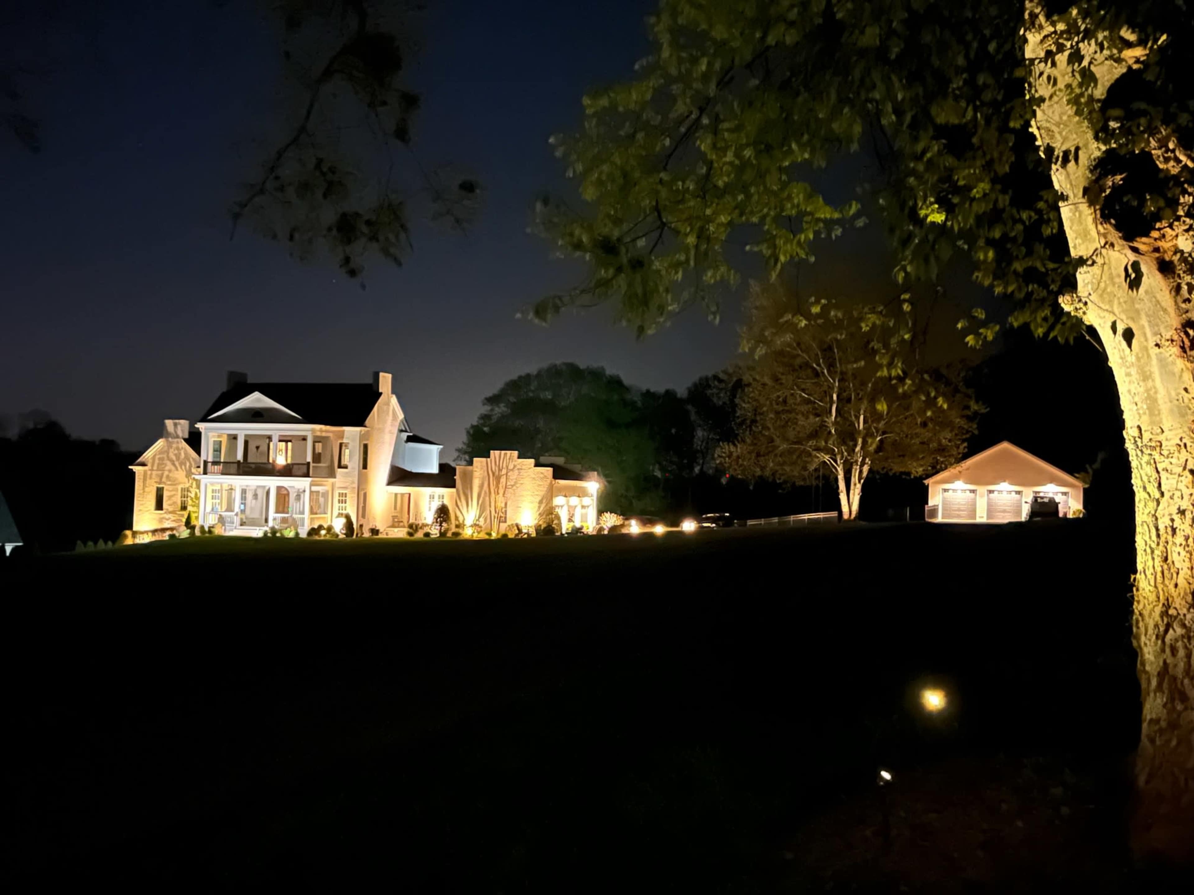 A well-lit house with a landscaped yard and a nearby garage is viewed at night.