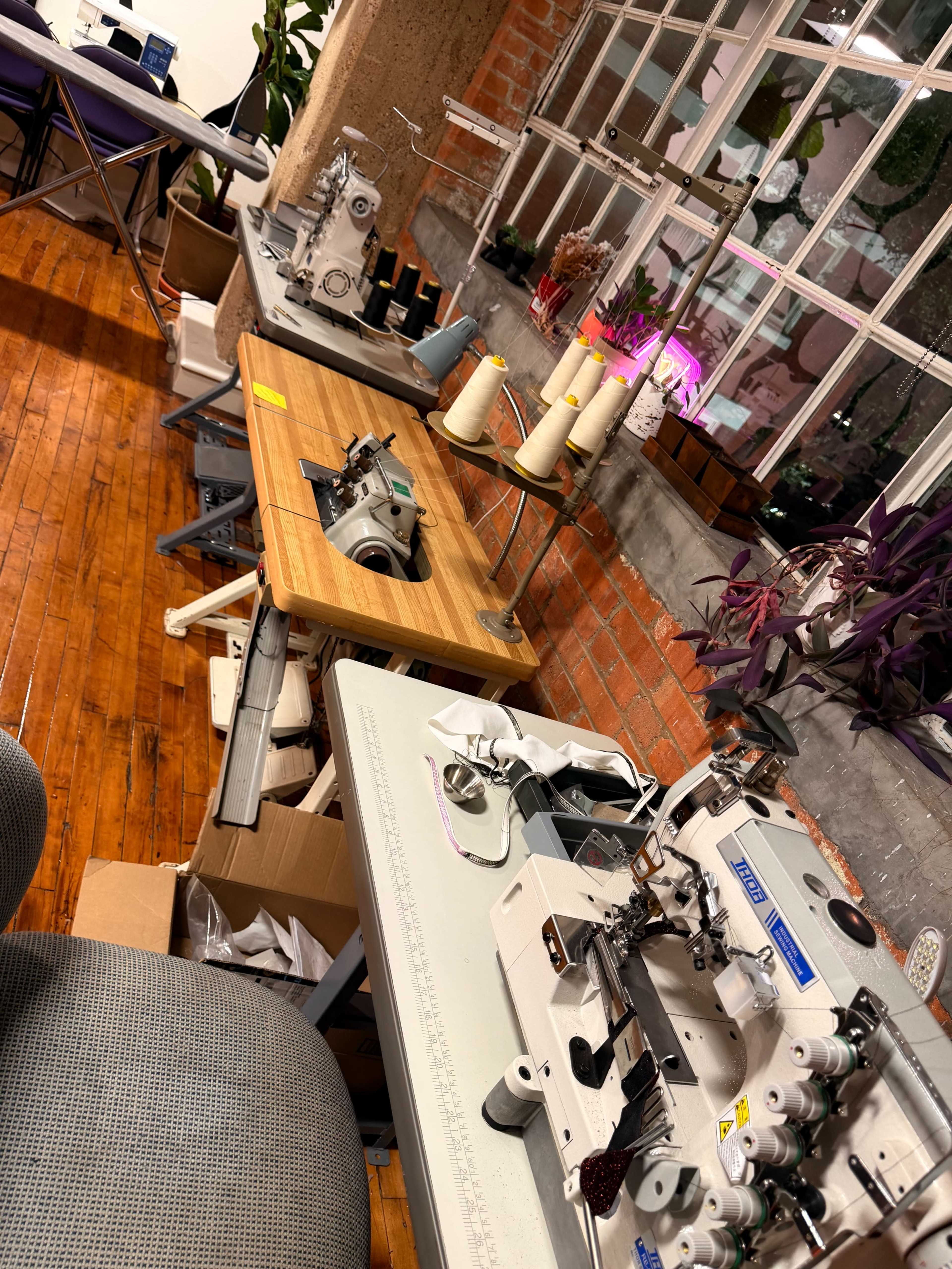 The image shows a workspace with several sewing machines and a wooden table amidst plants and a large window.