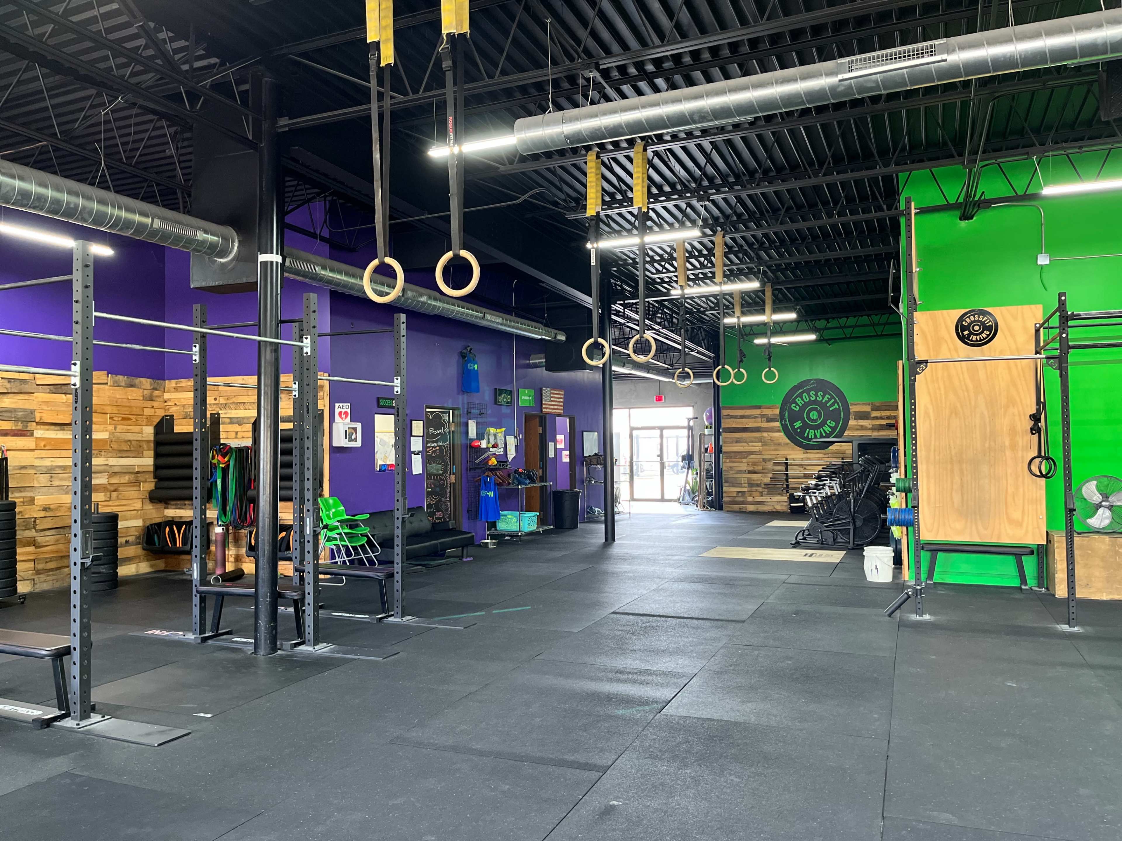 An indoor gym with a large open space, featuring workout equipment, hanging rings, and vibrant purple and green walls.