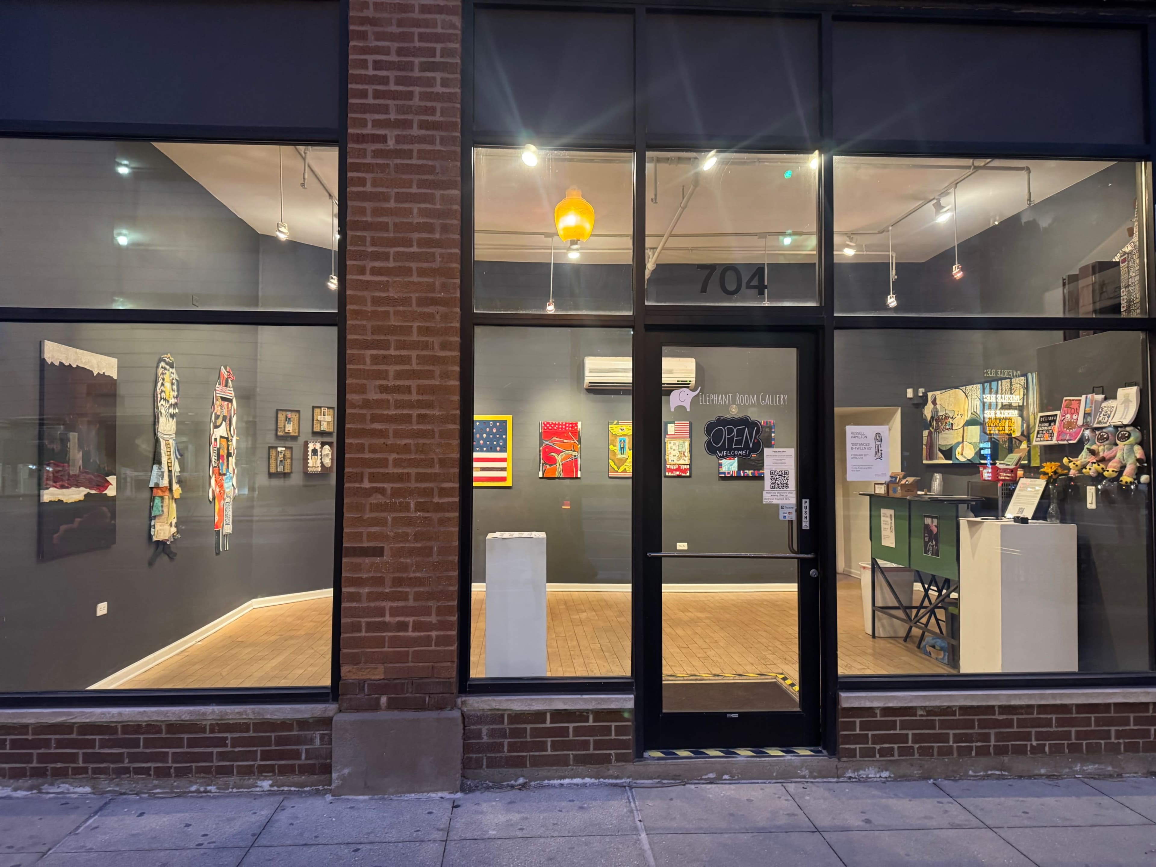 The image shows the exterior of an art gallery with large windows displaying various artworks and an "OPEN" sign.
