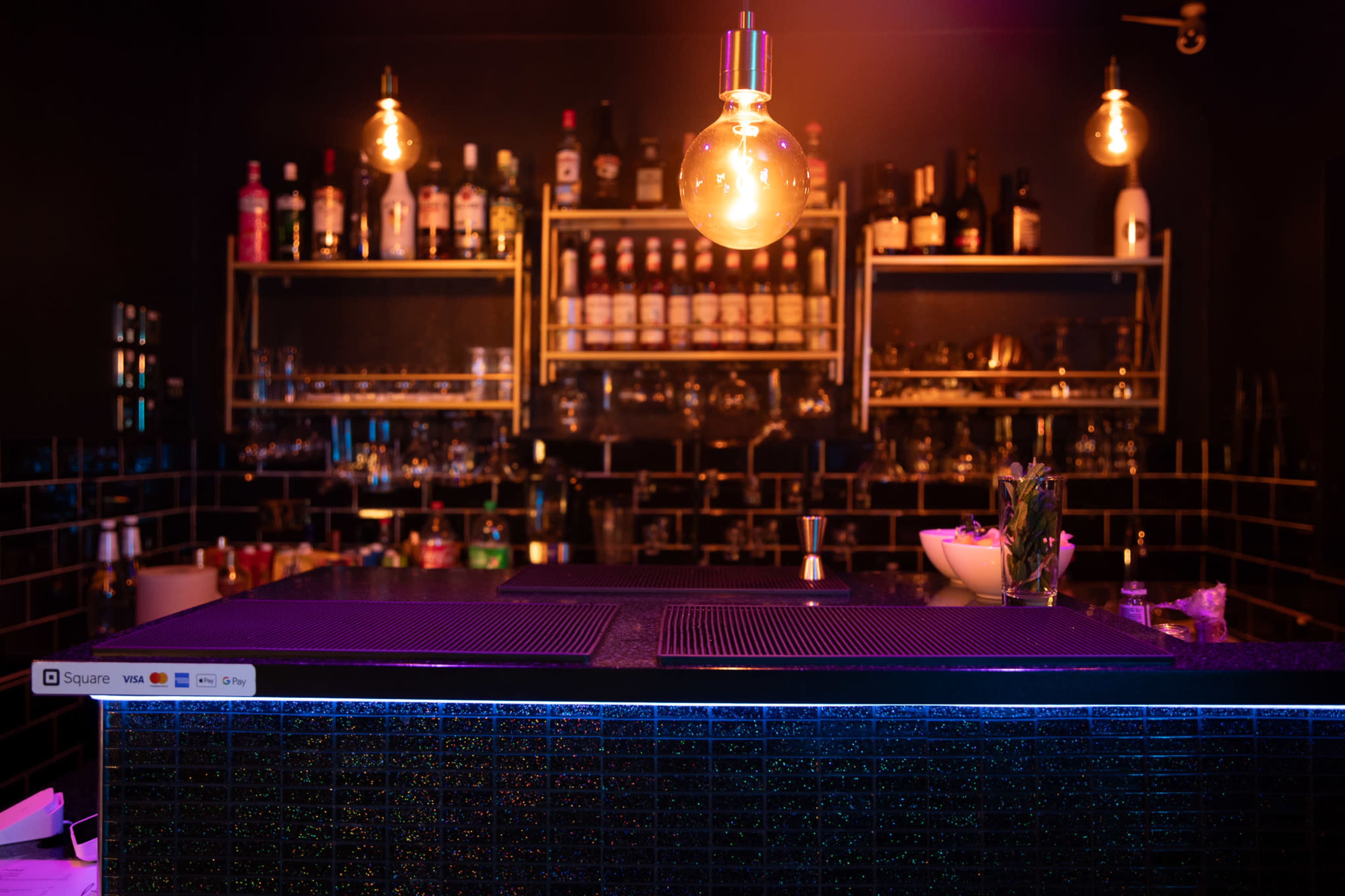 The image shows a dimly lit modern bar with a variety of liquor bottles displayed on shelves behind the counter.