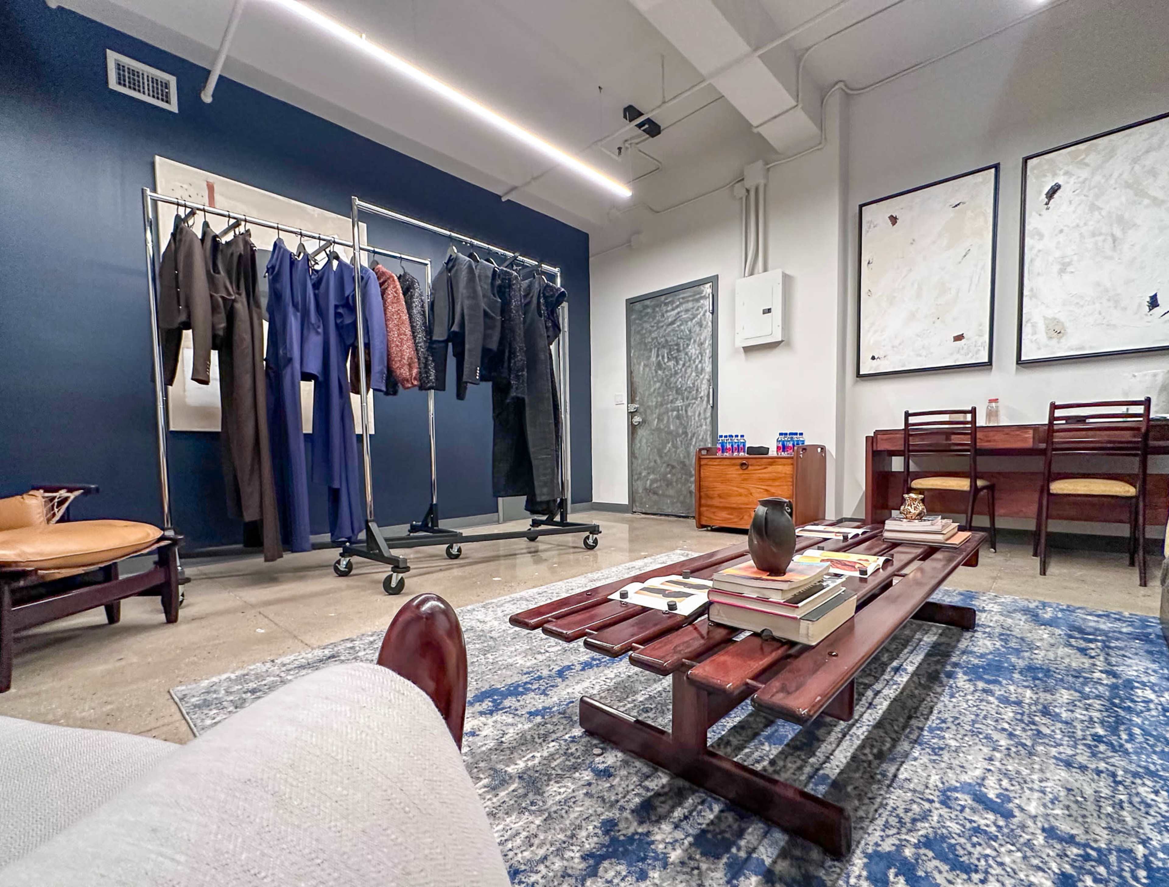 The image shows a contemporary interior space featuring a clothing rack with various garments, a coffee table with books, and a few chairs arranged around a patterned rug.