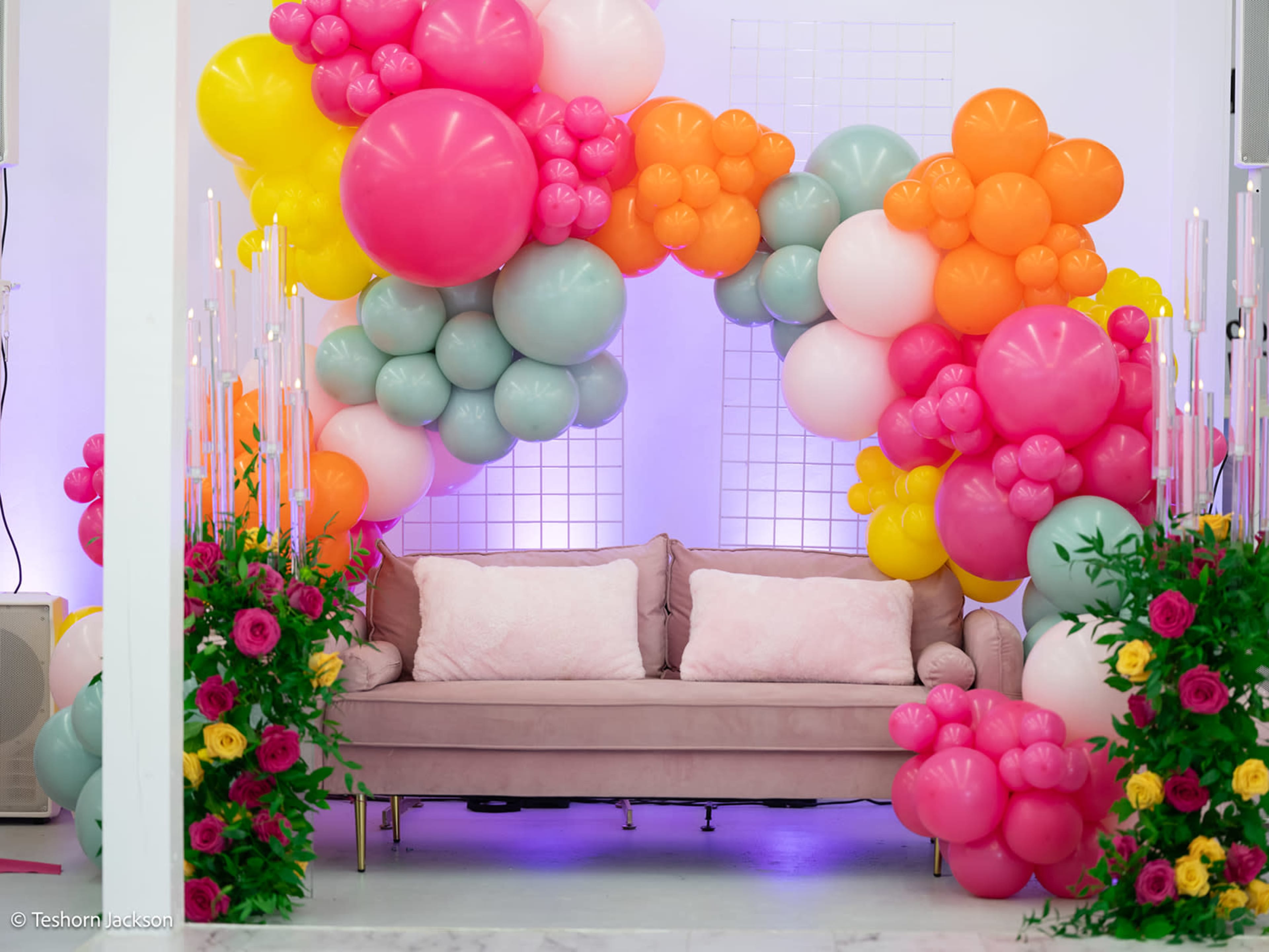 A sofa is positioned under a large arch of colorful balloons, surrounded by floral arrangements.