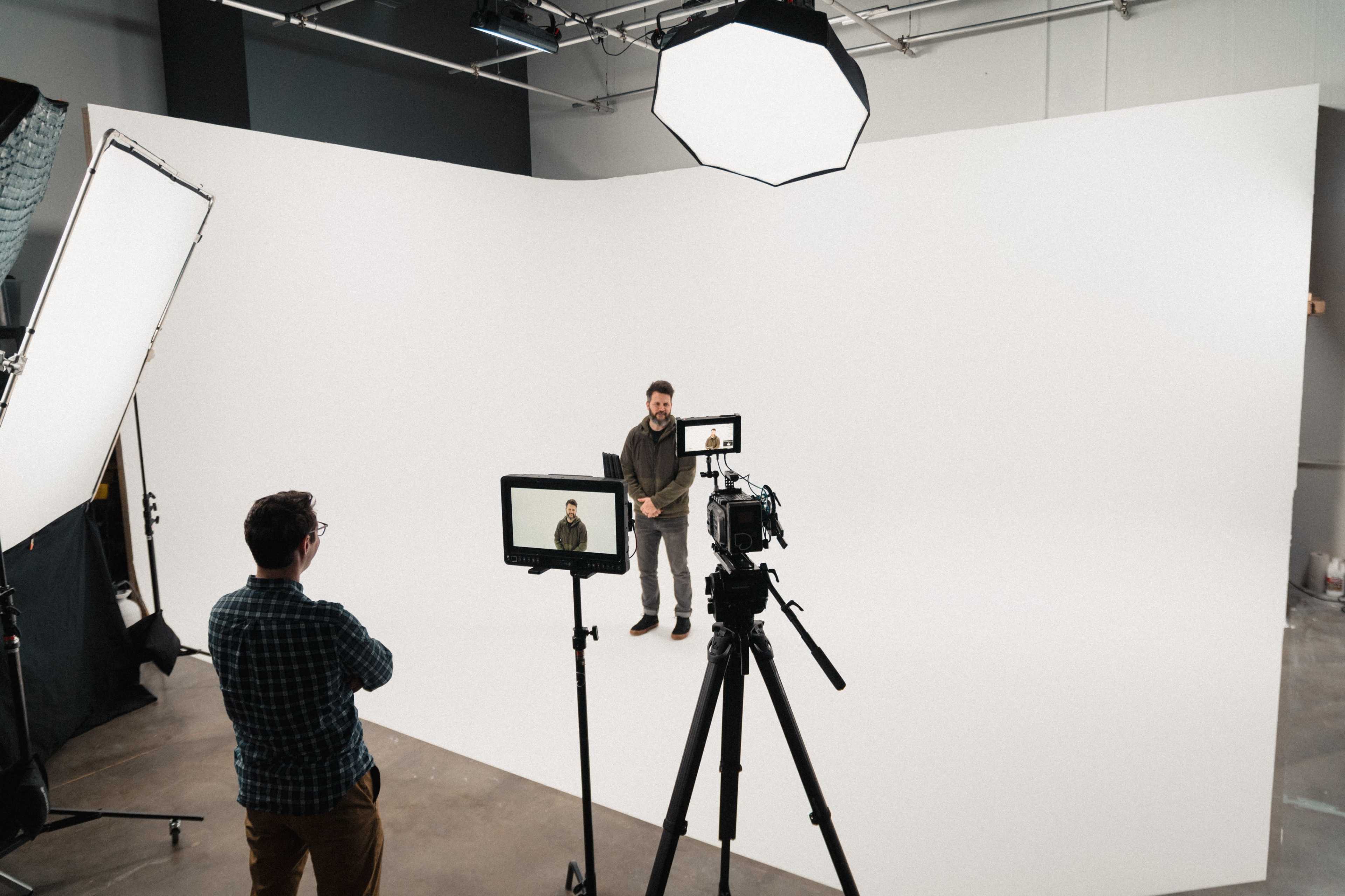 A person stands in front of a white backdrop while another individual operates a camera on a tripod.
