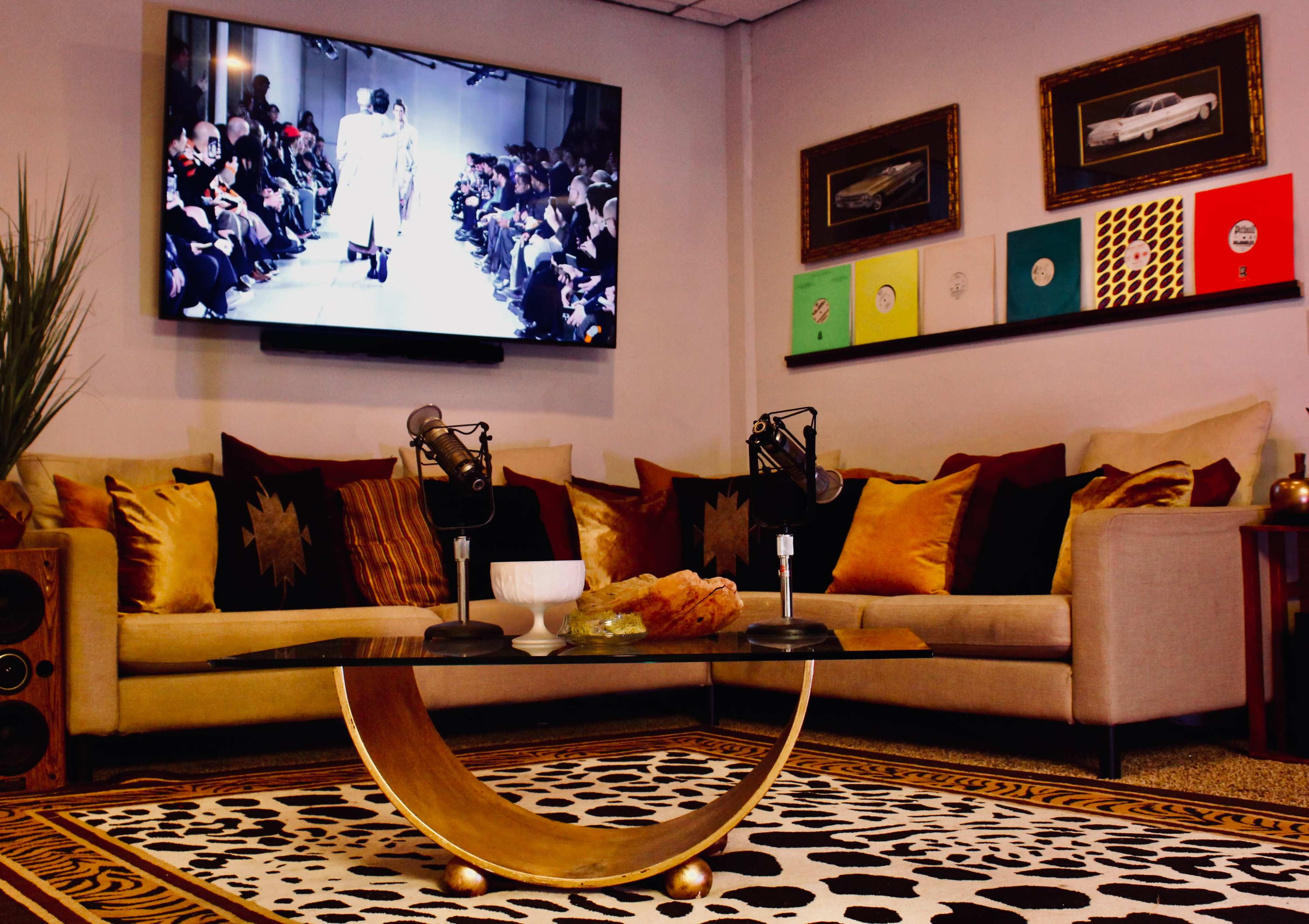 A stylish lounge area with a cream-colored sofa, a glass coffee table, and two microphones, while a large screen displays a fashion show.