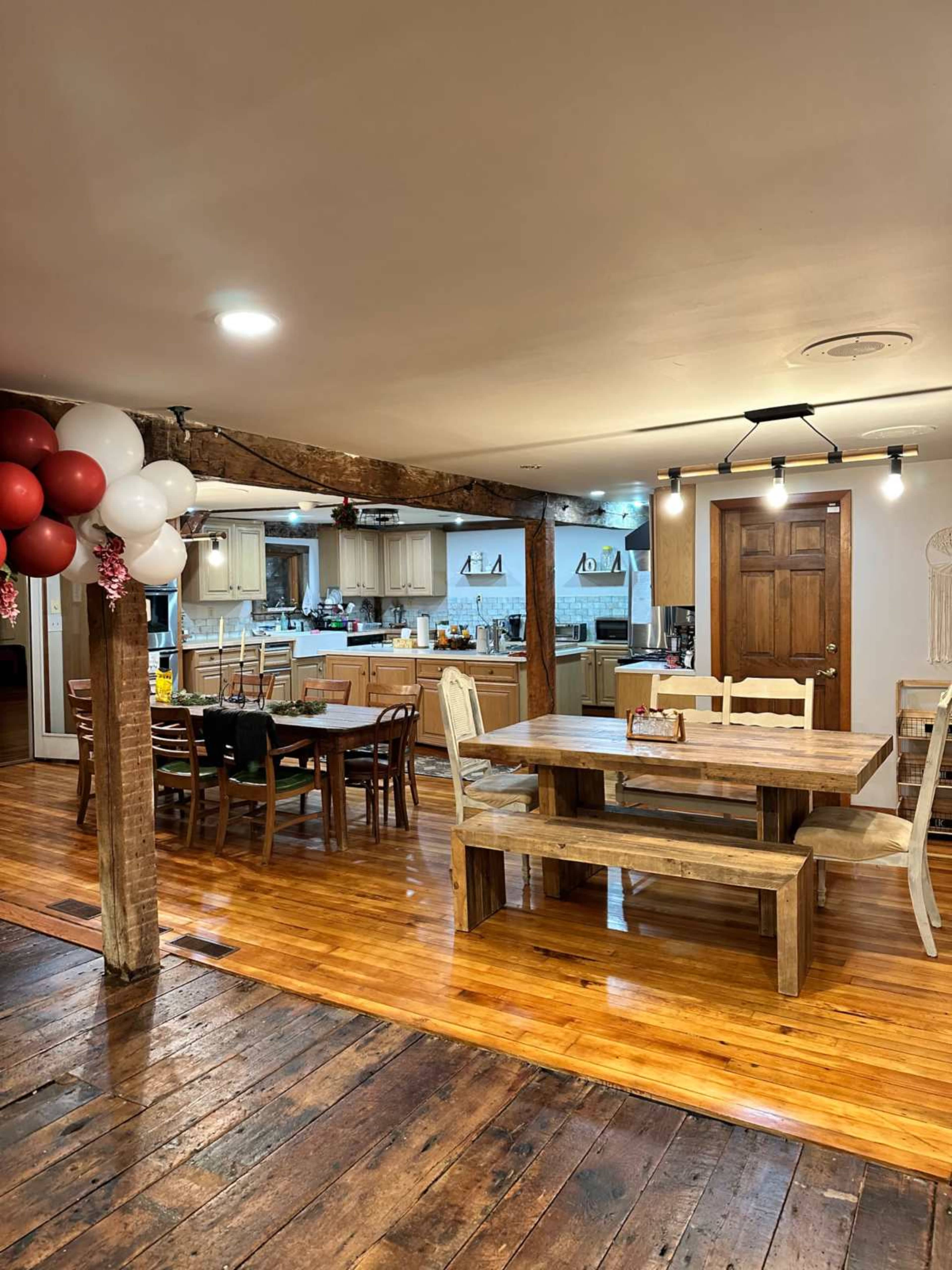 The image shows a spacious dining area with a wooden table and chairs, leading into a kitchen with modern appliances and wooden cabinetry.