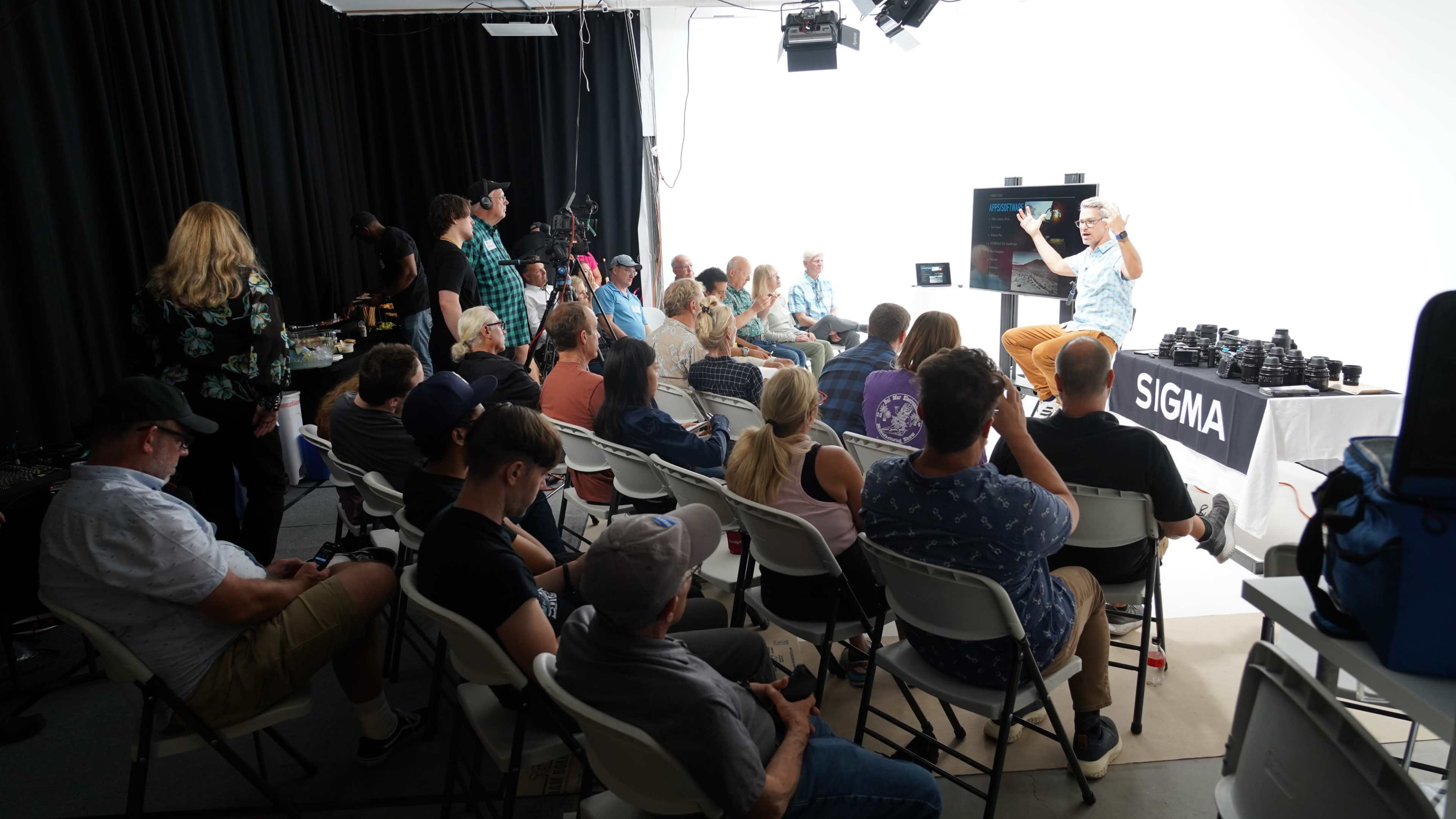 A group of people is seated in a studio, watching a presentation being given by a speaker in front of a light backdrop.