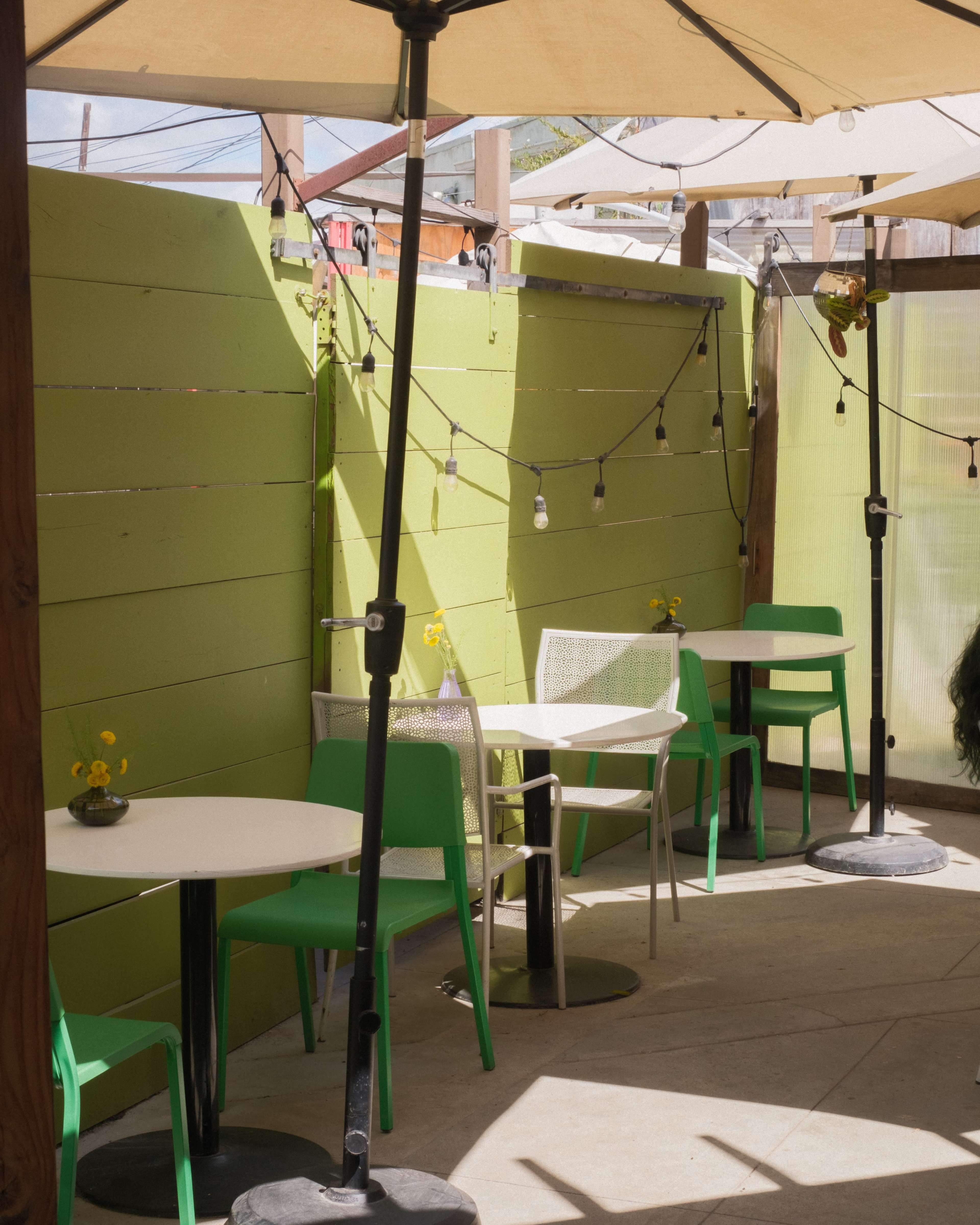 The image shows a small outdoor seating area with green walls, two tables, and chairs under large umbrellas.