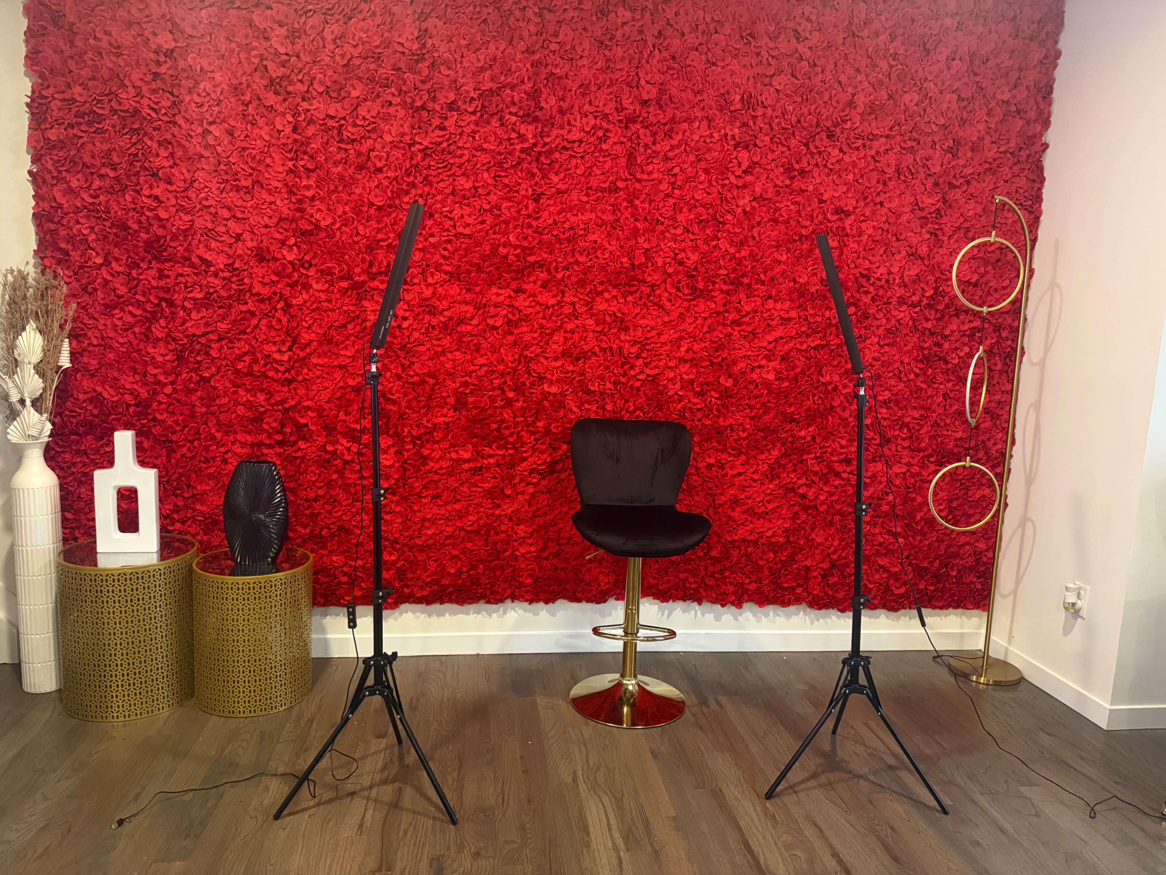 The image shows a room with a vibrant red floral wall, a black barstool sitting on a wooden floor, and two lighting stands positioned on either side.