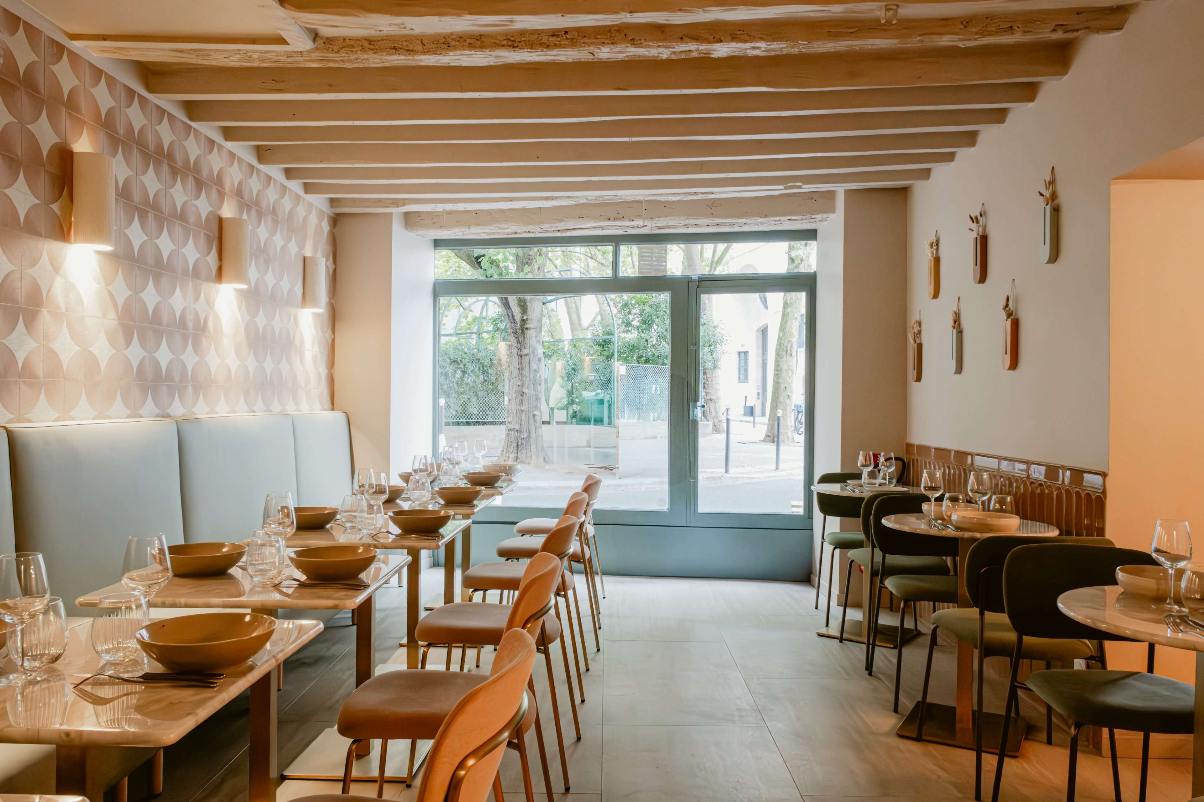 The image shows a modern restaurant interior featuring wooden beams, neatly arranged tables with place settings, and large windows providing natural light.