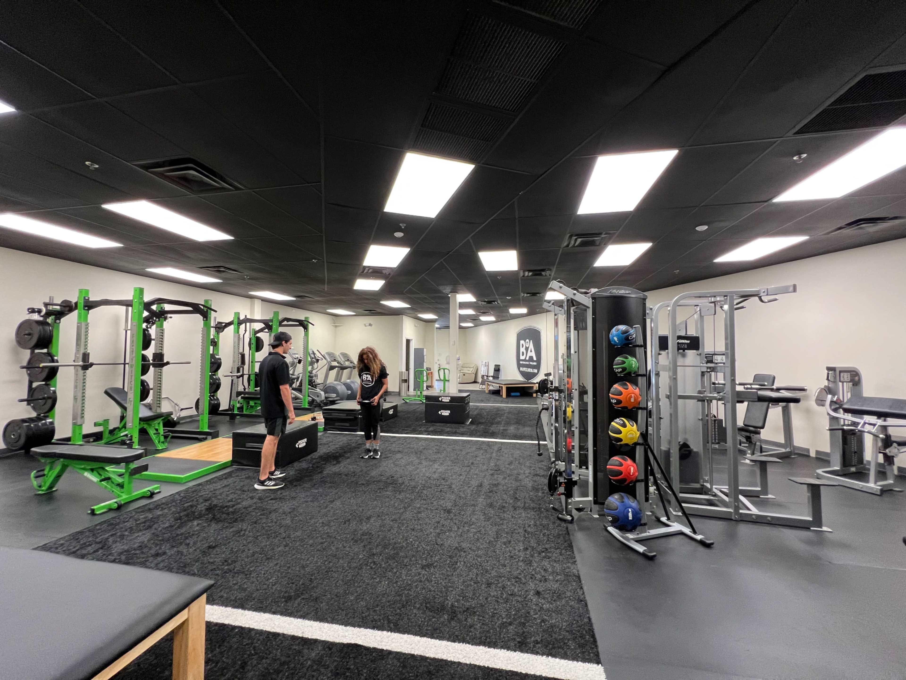 A modern gym features various exercise equipment, including weights and cardio machines, with two individuals engaged in conversation.