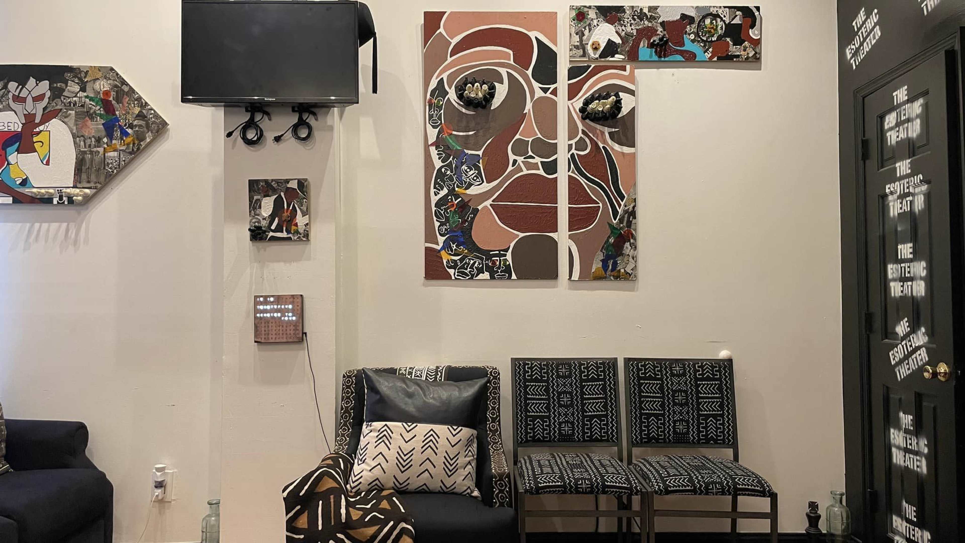 A modern indoor space featuring a television mounted on the wall, several pieces of artistic wall decor, and a seating area with patterned chairs and cushions.