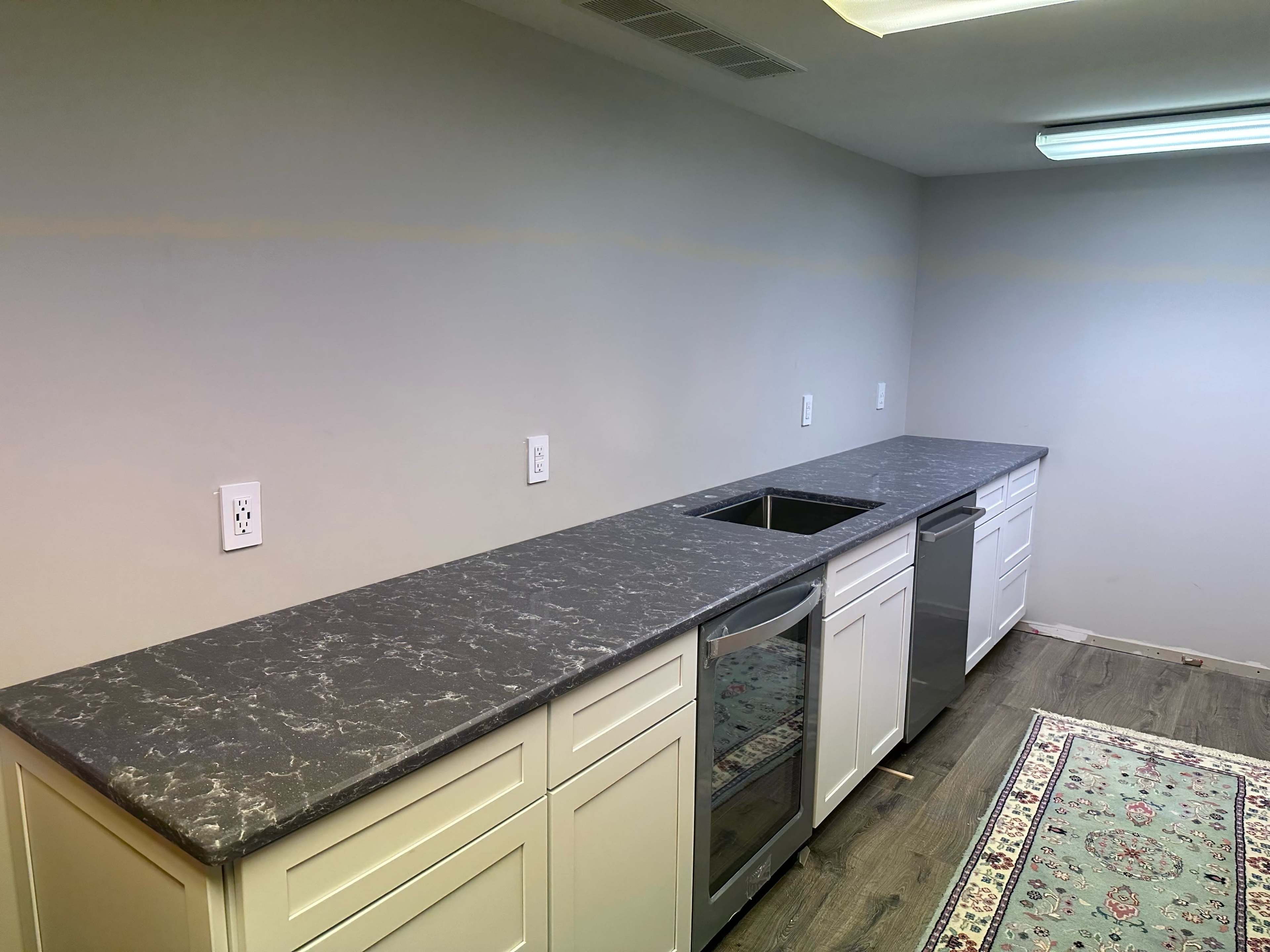 The image shows a kitchen with a long countertop, white cabinets, and a gray surface, alongside a rug on the floor.