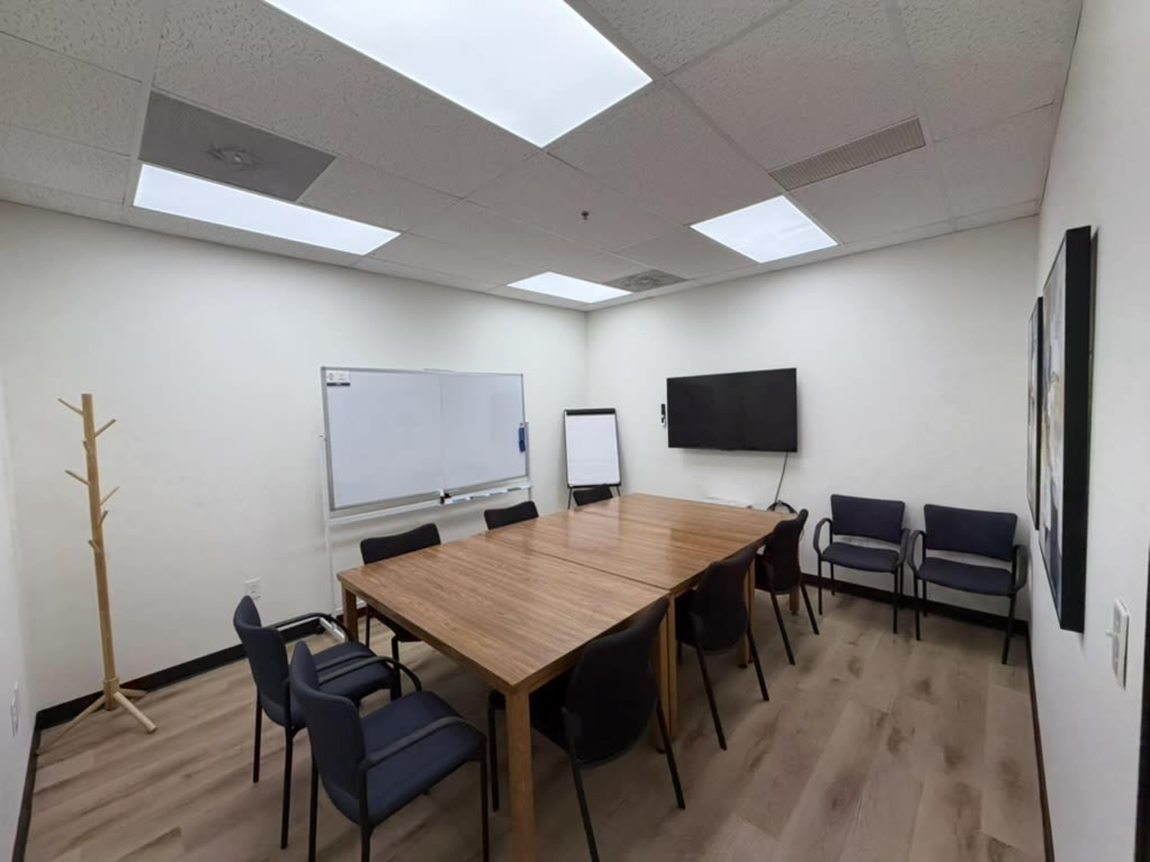 A small conference room features a large wooden table surrounded by eight chairs, a whiteboard, and a wall-mounted television.