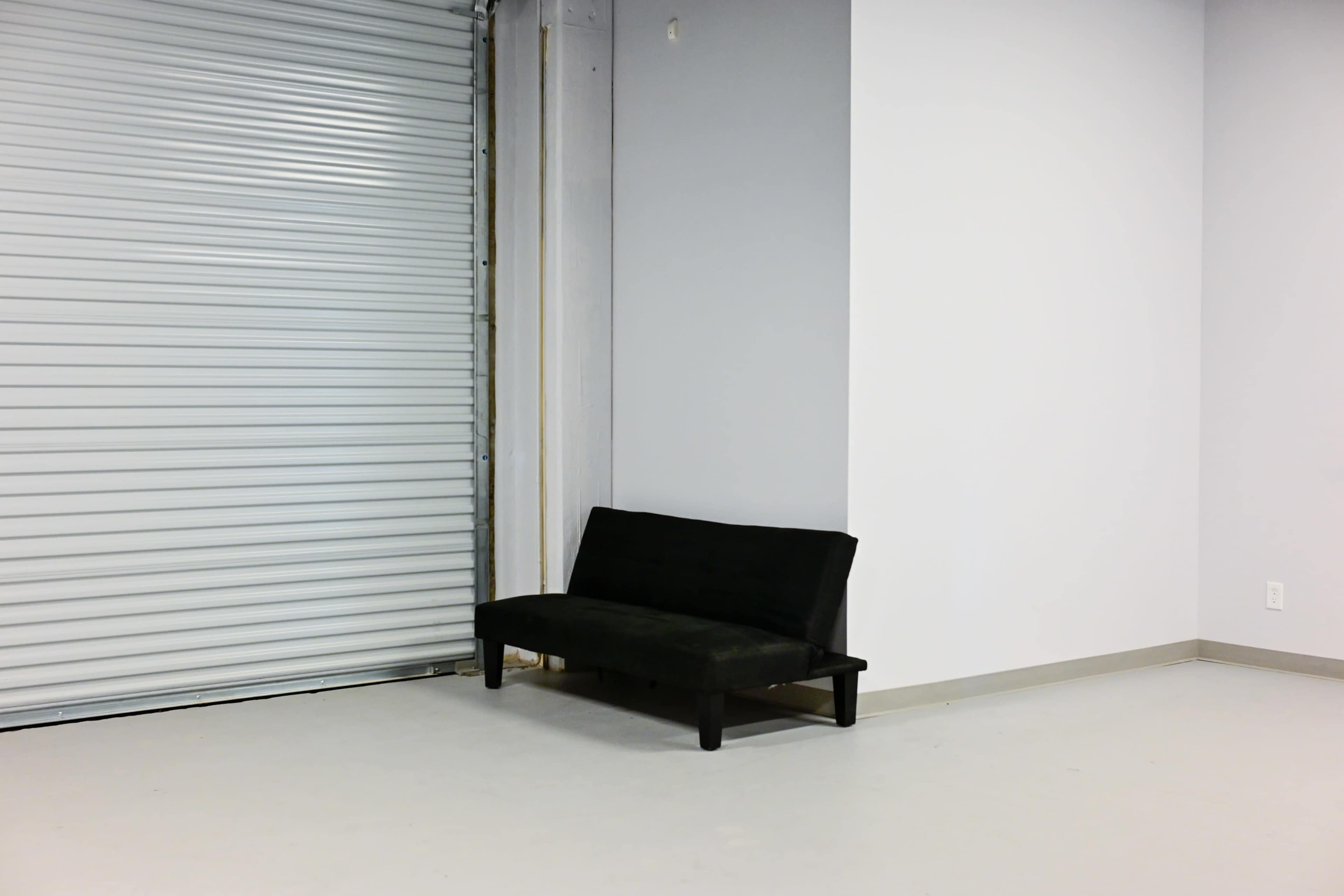 A black futon is positioned against a plain white wall next to a rolled-up metal garage door.