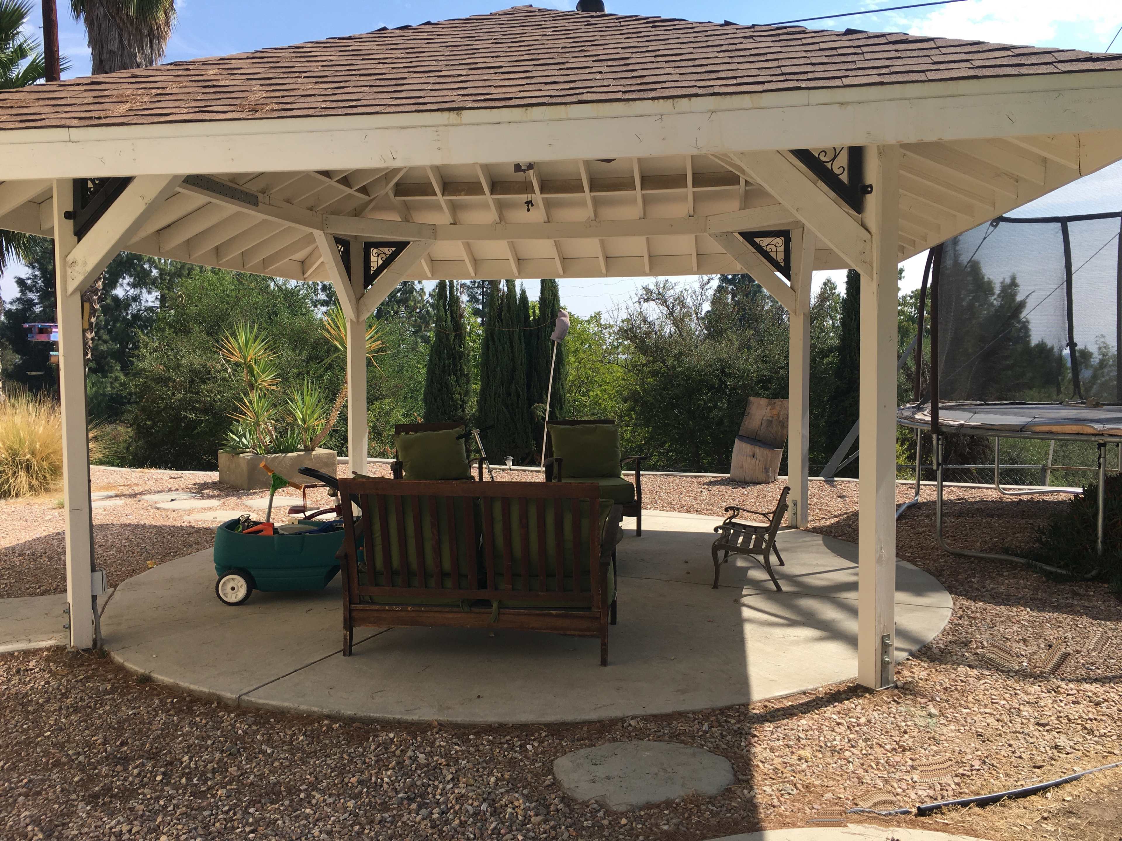 A gazebo with a wooden bench and two green chairs overlooks a landscaped area with a trampoline in the background.