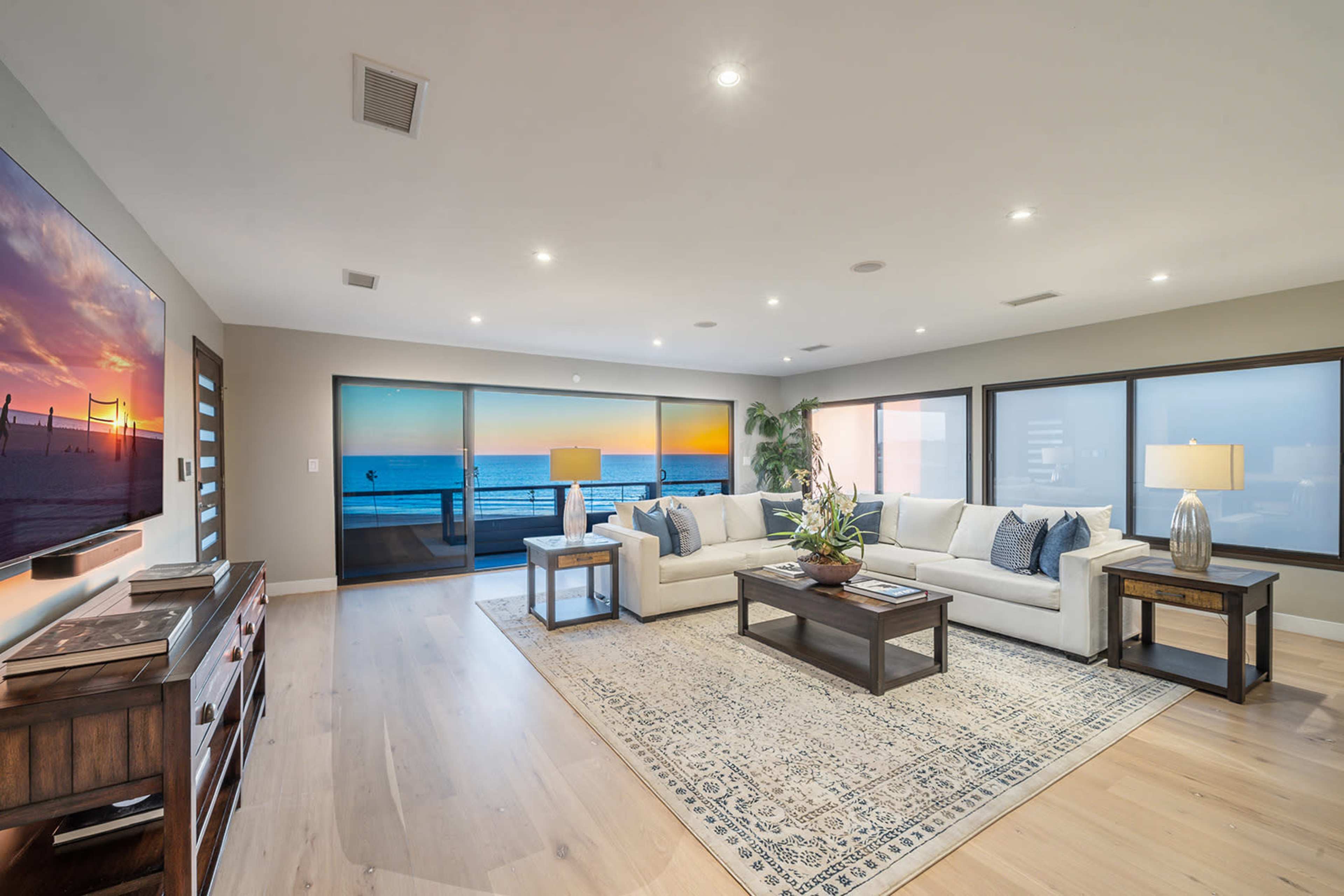 A modern living room features a large sectional sofa, a coffee table, and a television, with expansive windows showcasing a sunset view over the ocean.
