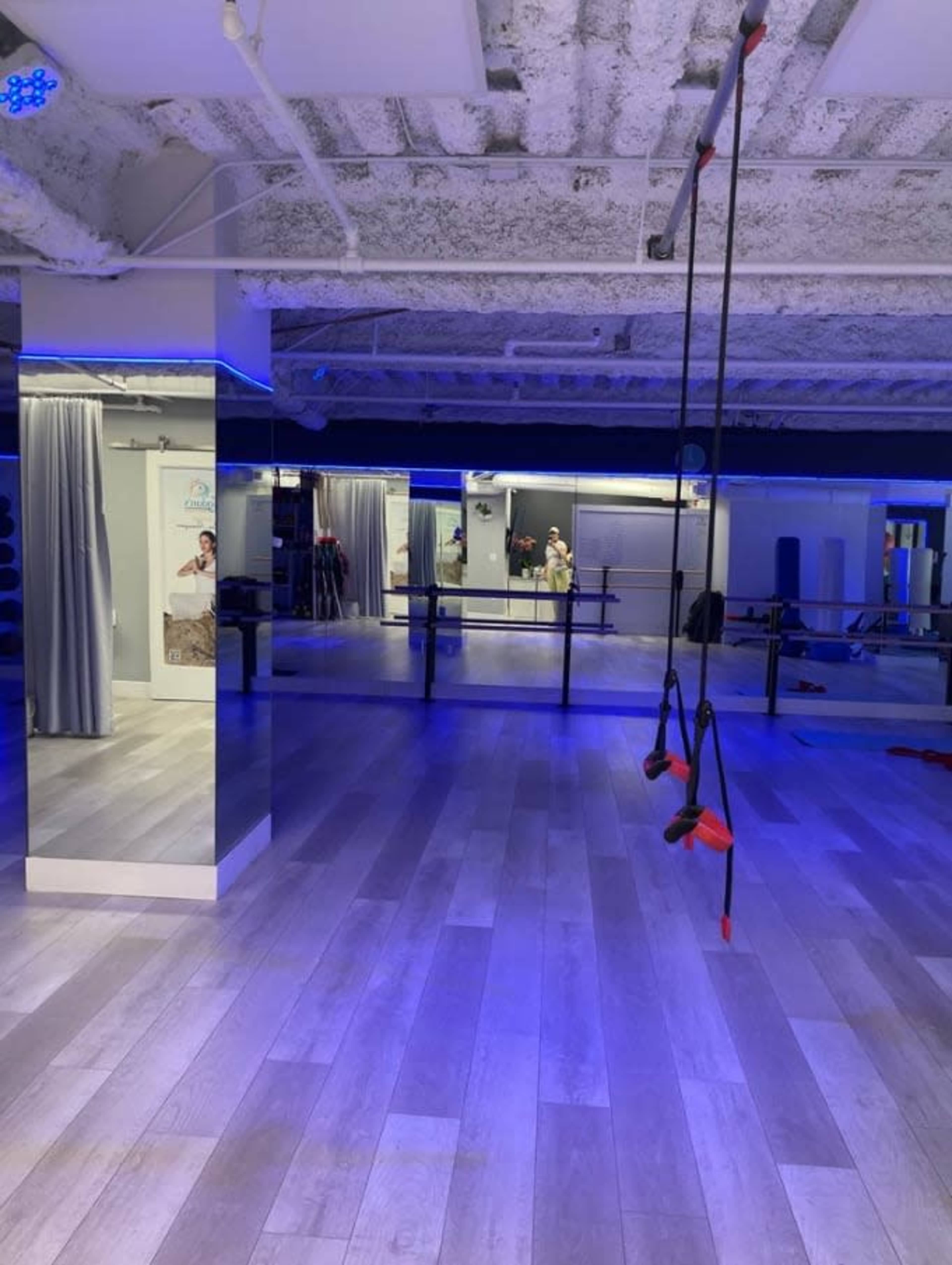 The image shows a spacious fitness studio with wooden flooring, large mirrors on one wall, and suspended exercise straps hanging from the ceiling under blue lighting.