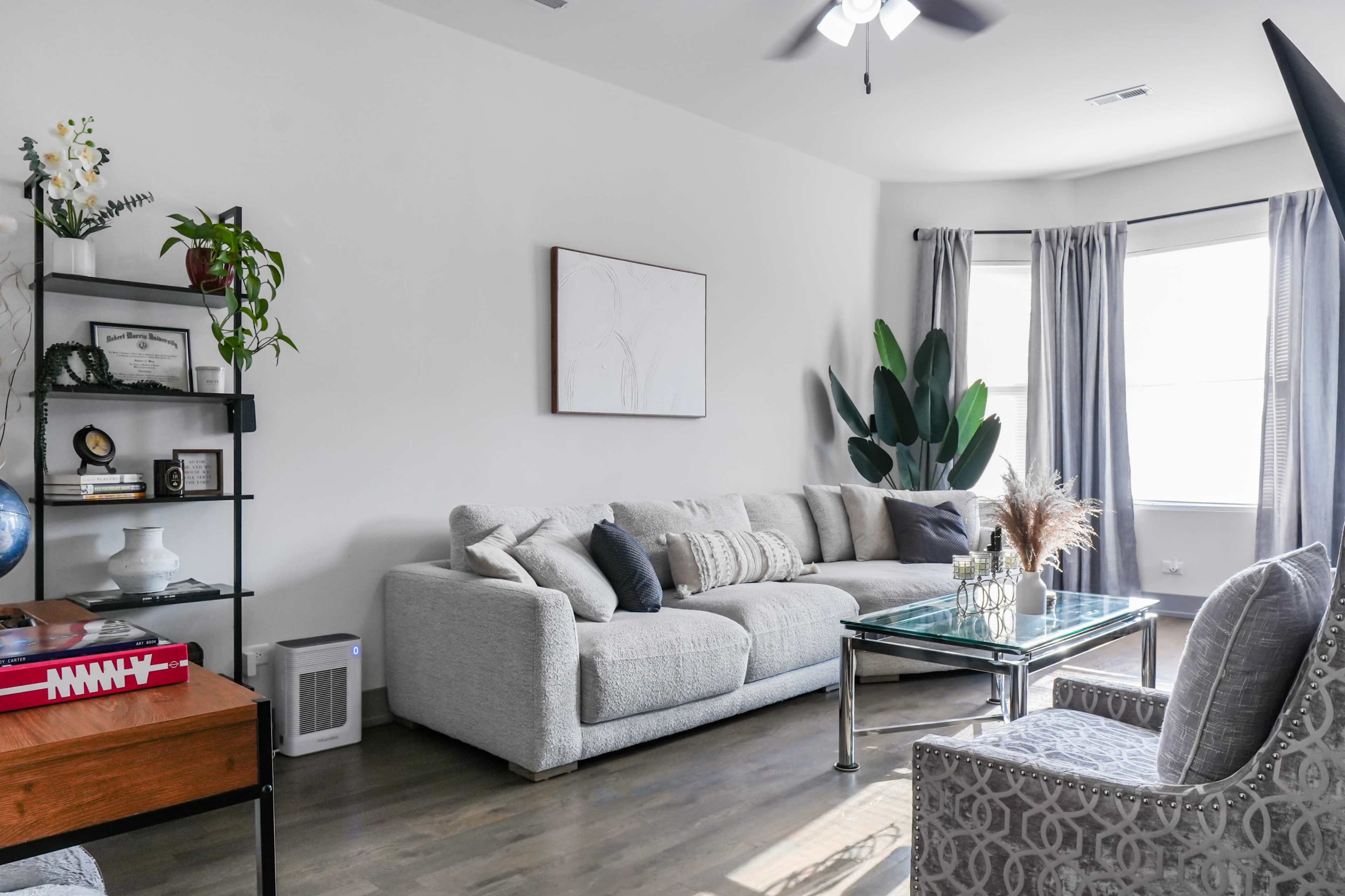 The image shows a modern living room featuring a light-colored sectional sofa, a glass coffee table, a potted plant, and a window with sheer curtains.