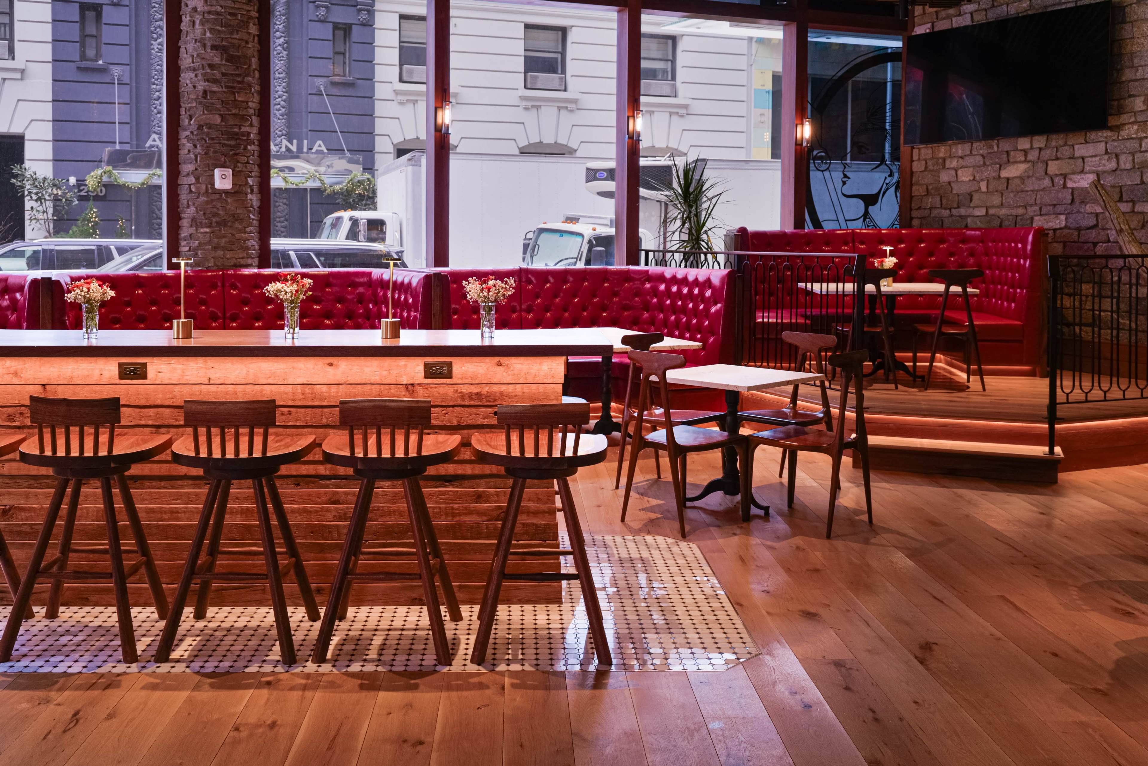 The image shows a stylish bar area with wooden stools, a polished wooden counter, and cozy seating along the windows adorned with potted plants.