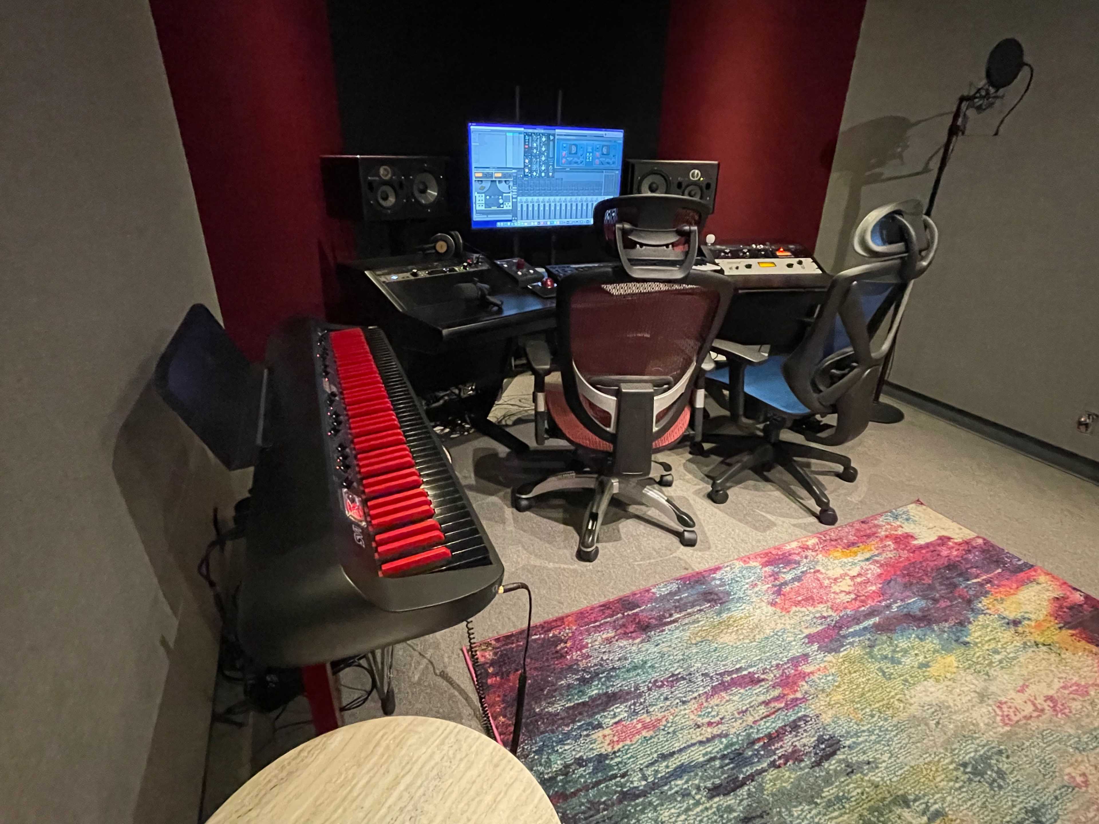 A recording studio with professional audio equipment, two chairs, a computer monitor displaying music software, and a keyboard positioned beside a rug.