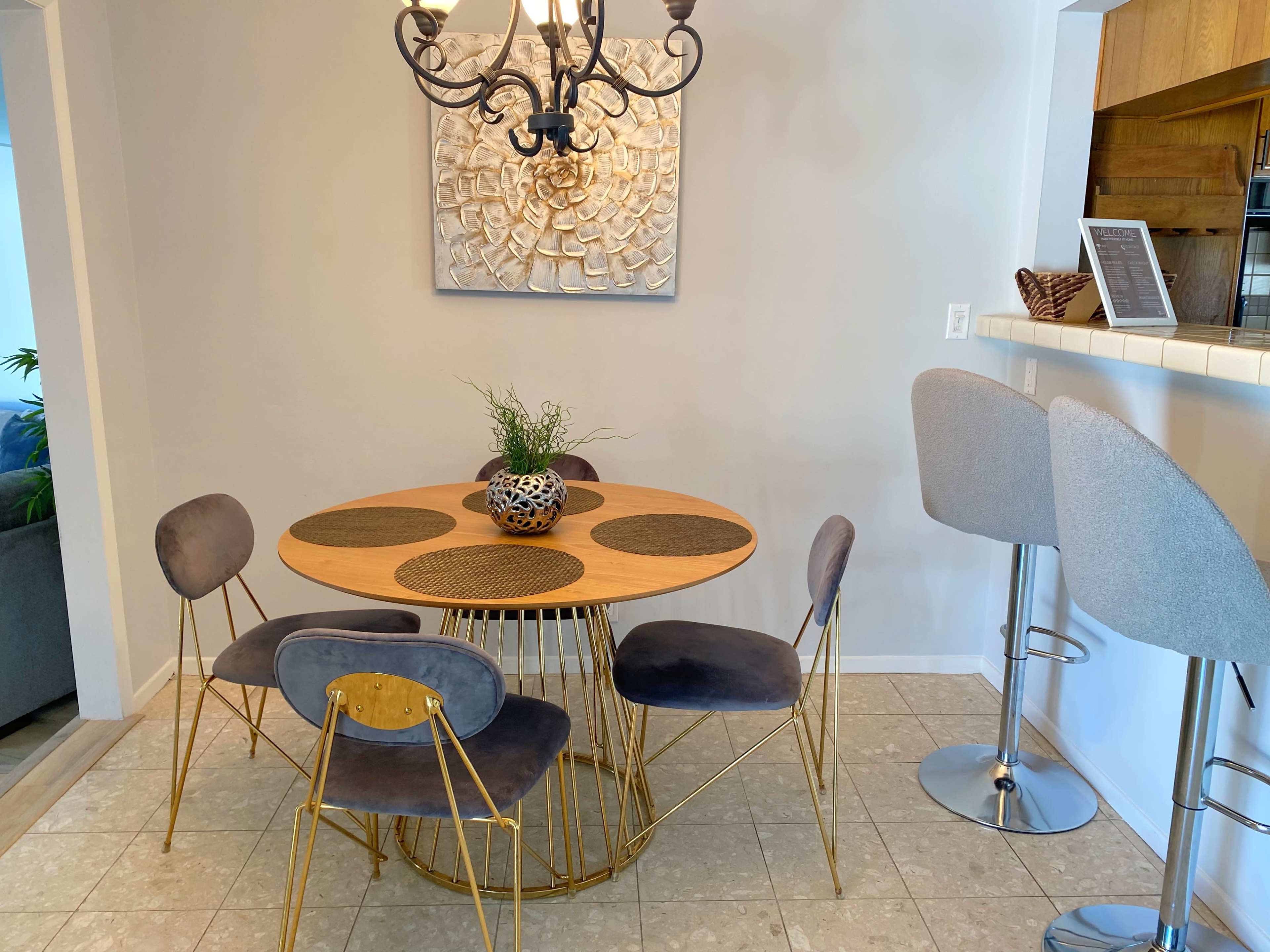 A round wooden dining table with gold legs and gray chairs is set in a well-lit dining area, accompanied by a simple decorative centerpiece and a wall-mounted art piece.