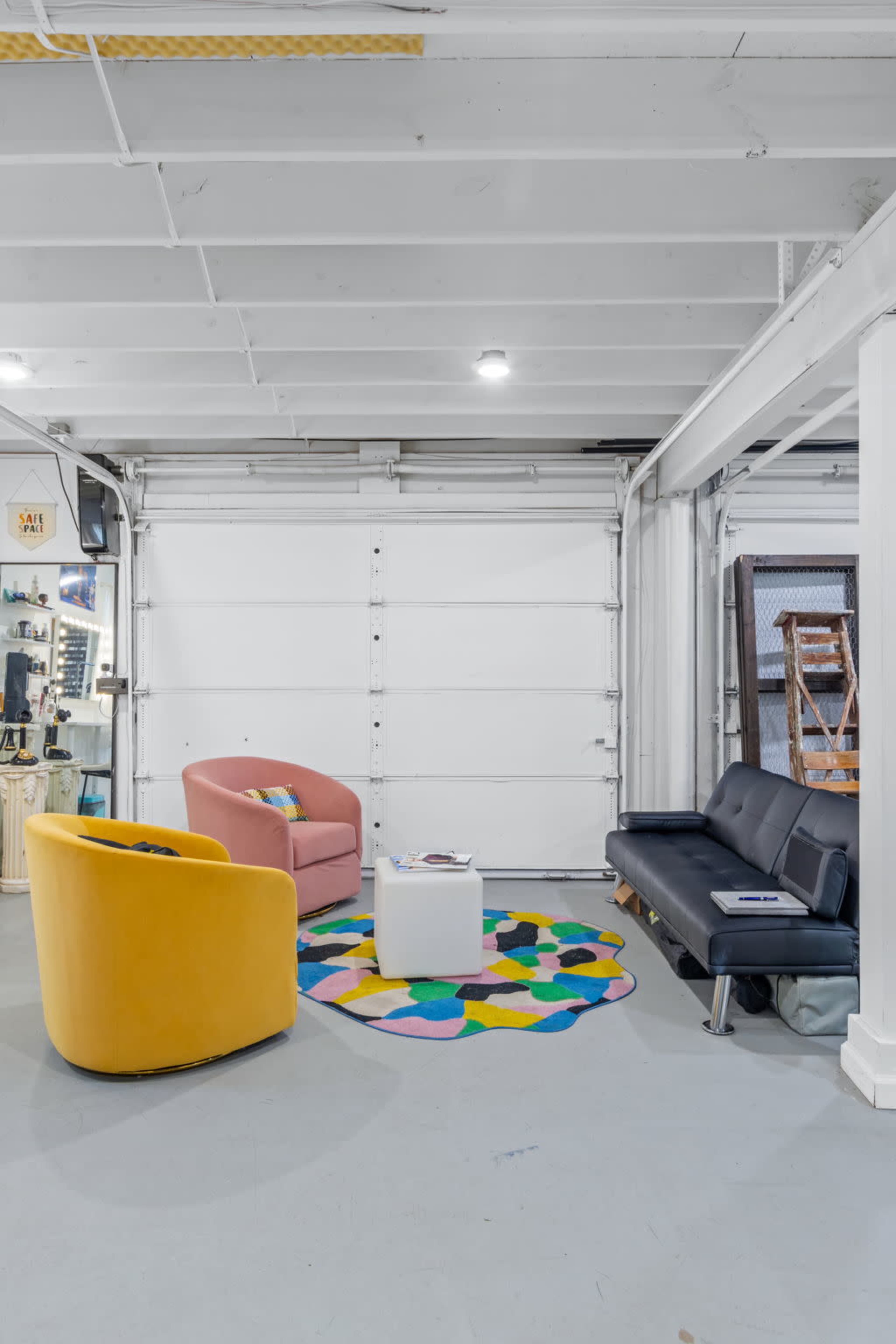 The image shows a modern garage space with a colorful area rug, two accent chairs in pink and yellow, and a black sofa facing a white coffee table.