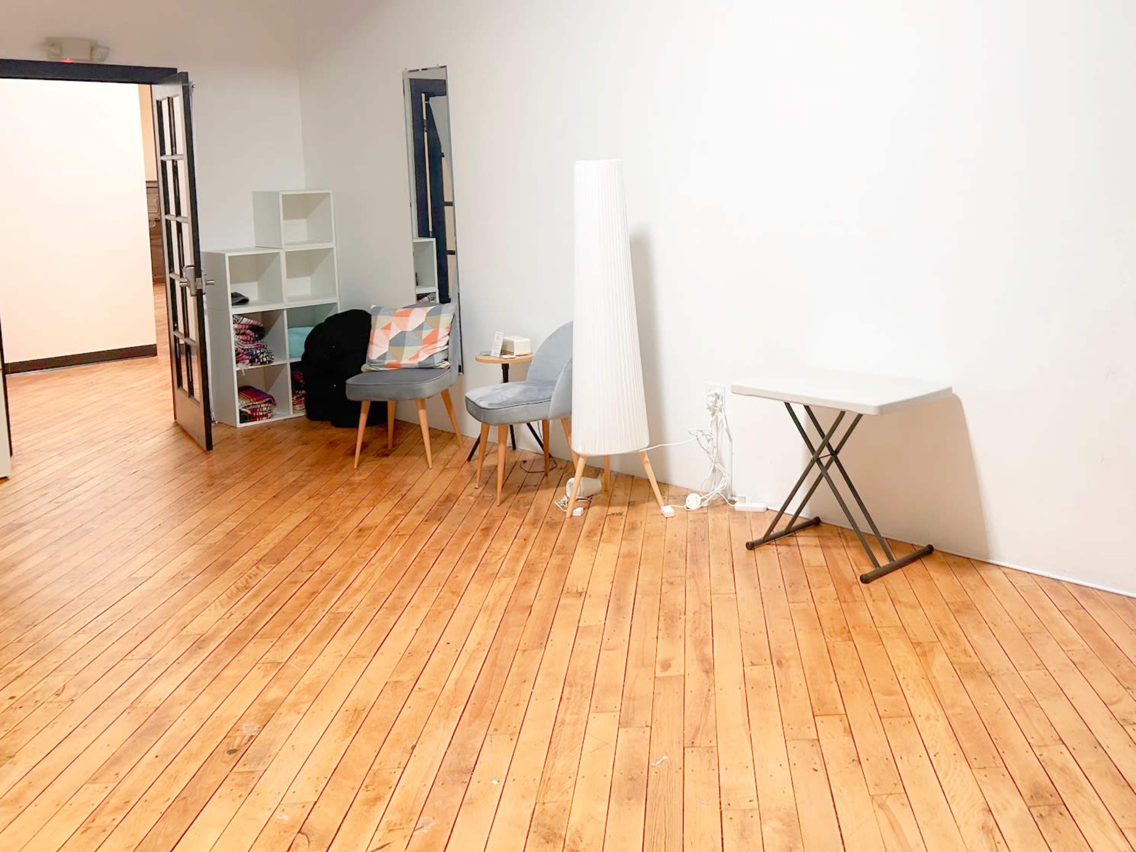 The room features wooden flooring with a door leading to another space, a light-colored floor lamp, two chairs, and a small table against a blank wall.