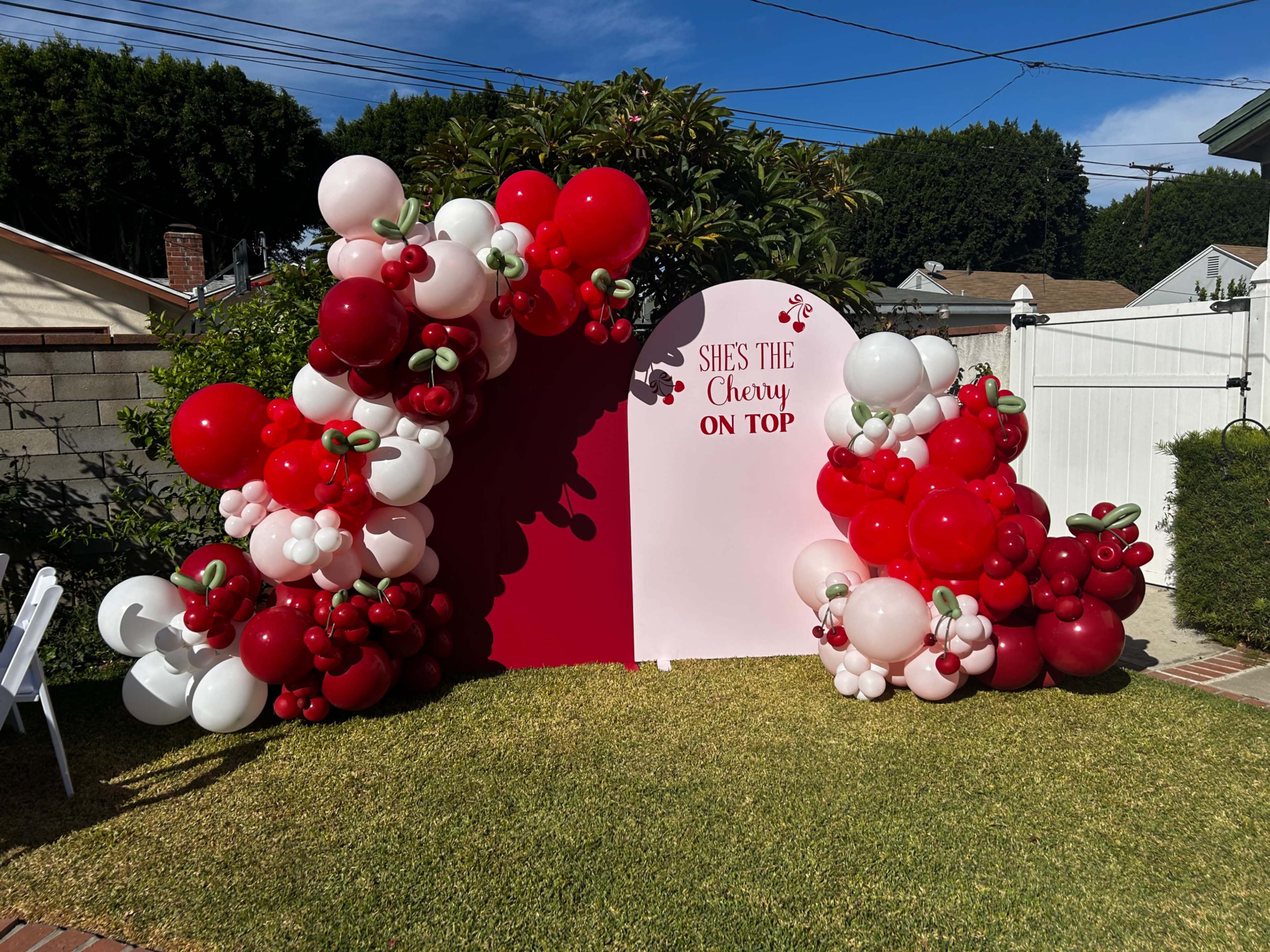 A colorful balloon arch featuring red, white, and pink balloons surrounds a pink backdrop with the phrase "SHE'S THE CHERRY ON TOP."