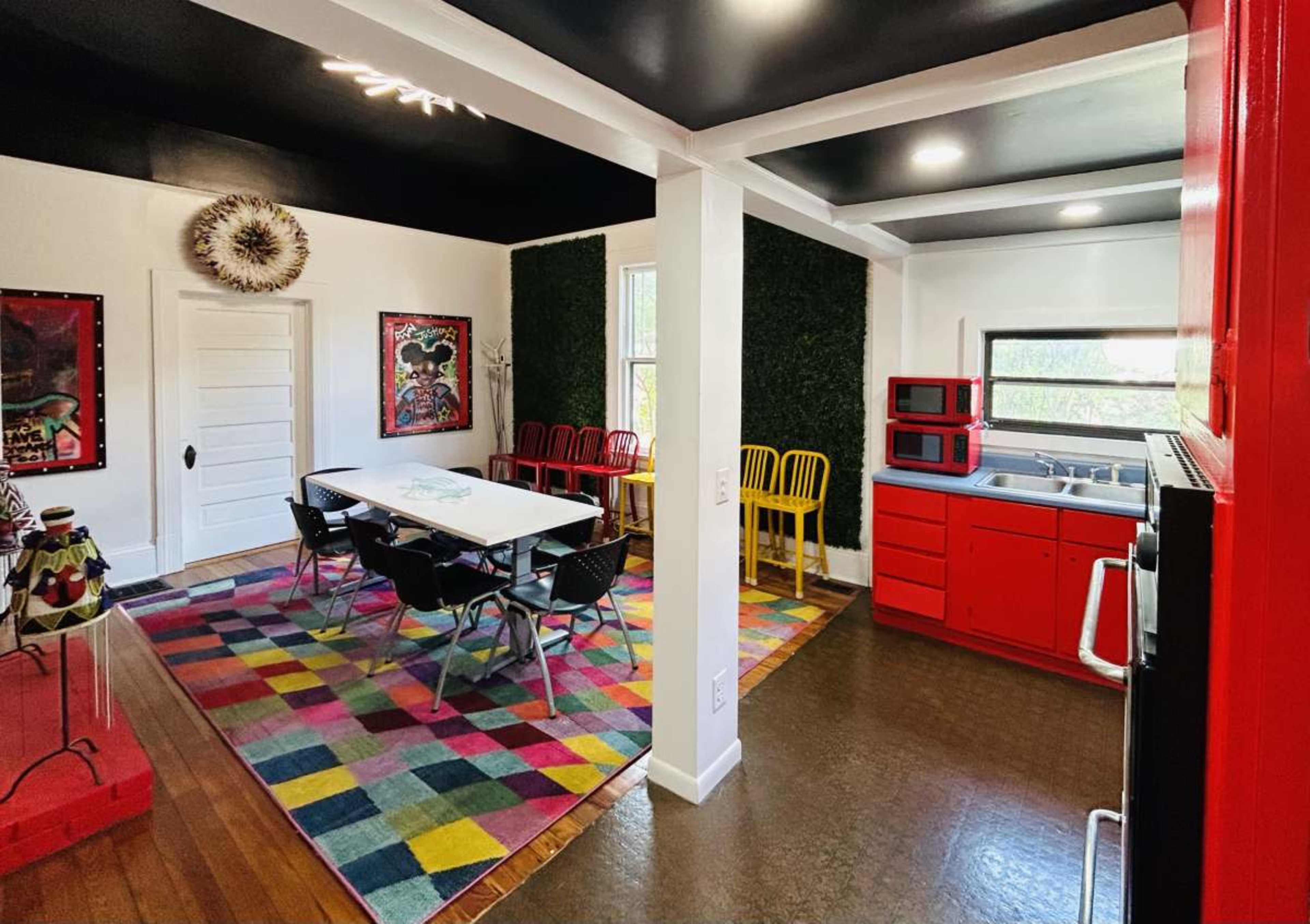 The image shows a contemporary kitchen and dining area featuring red cabinetry, a large table with black chairs, and colorful artwork on the walls.