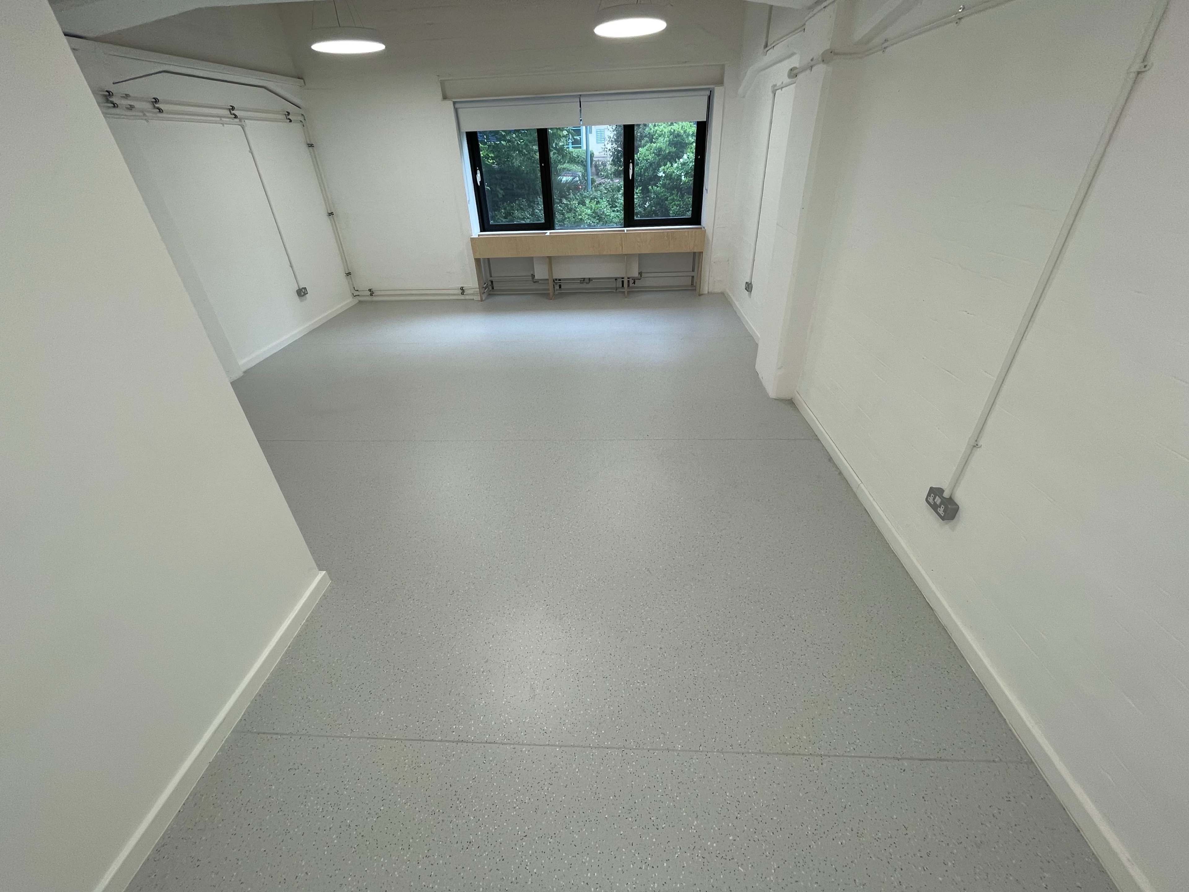 A spacious, empty room with a light gray floor and large windows overlooking greenery.