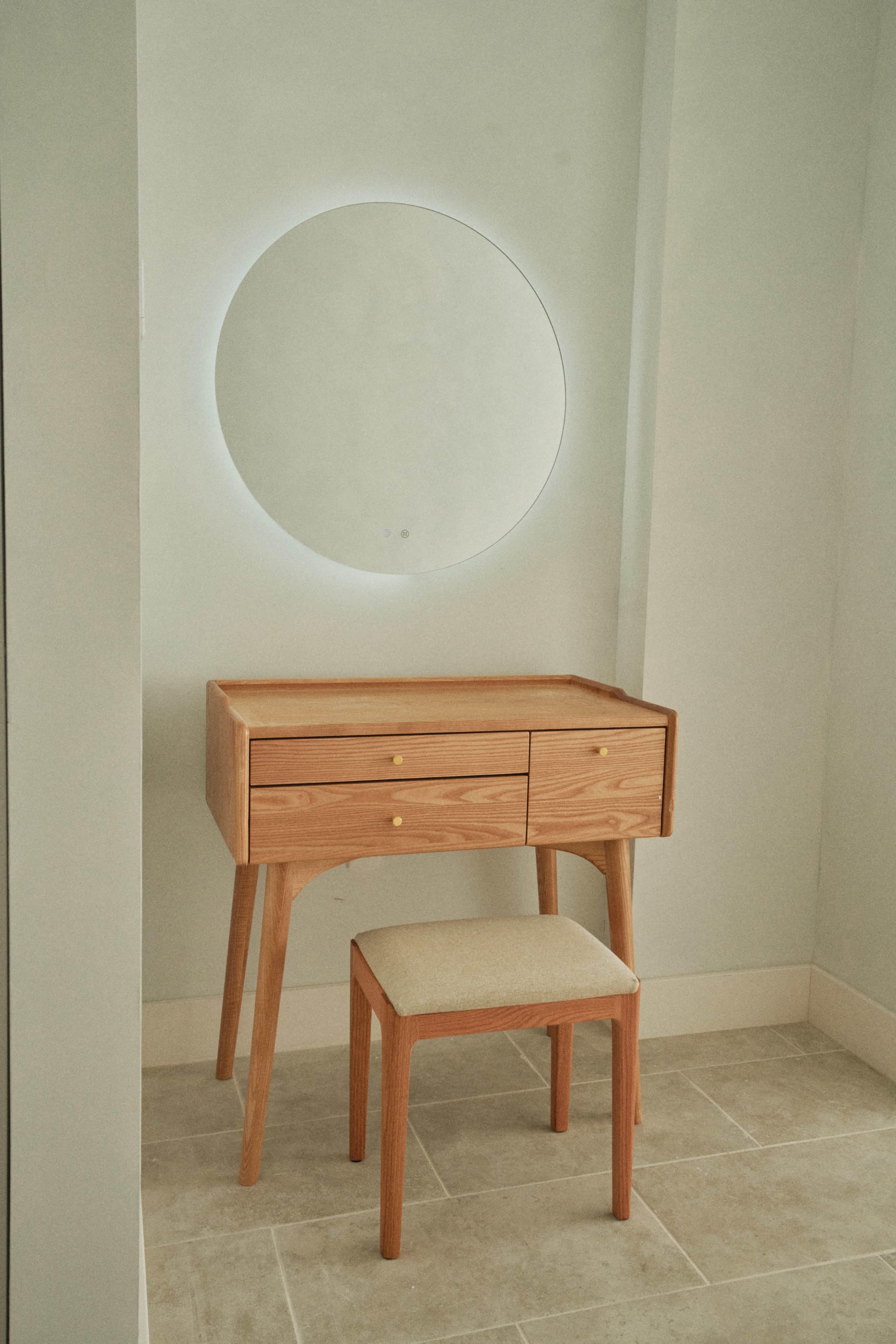 A wooden vanity with two drawers and a beige stool stands against a light gray wall, accompanied by a circular mirror above it.
