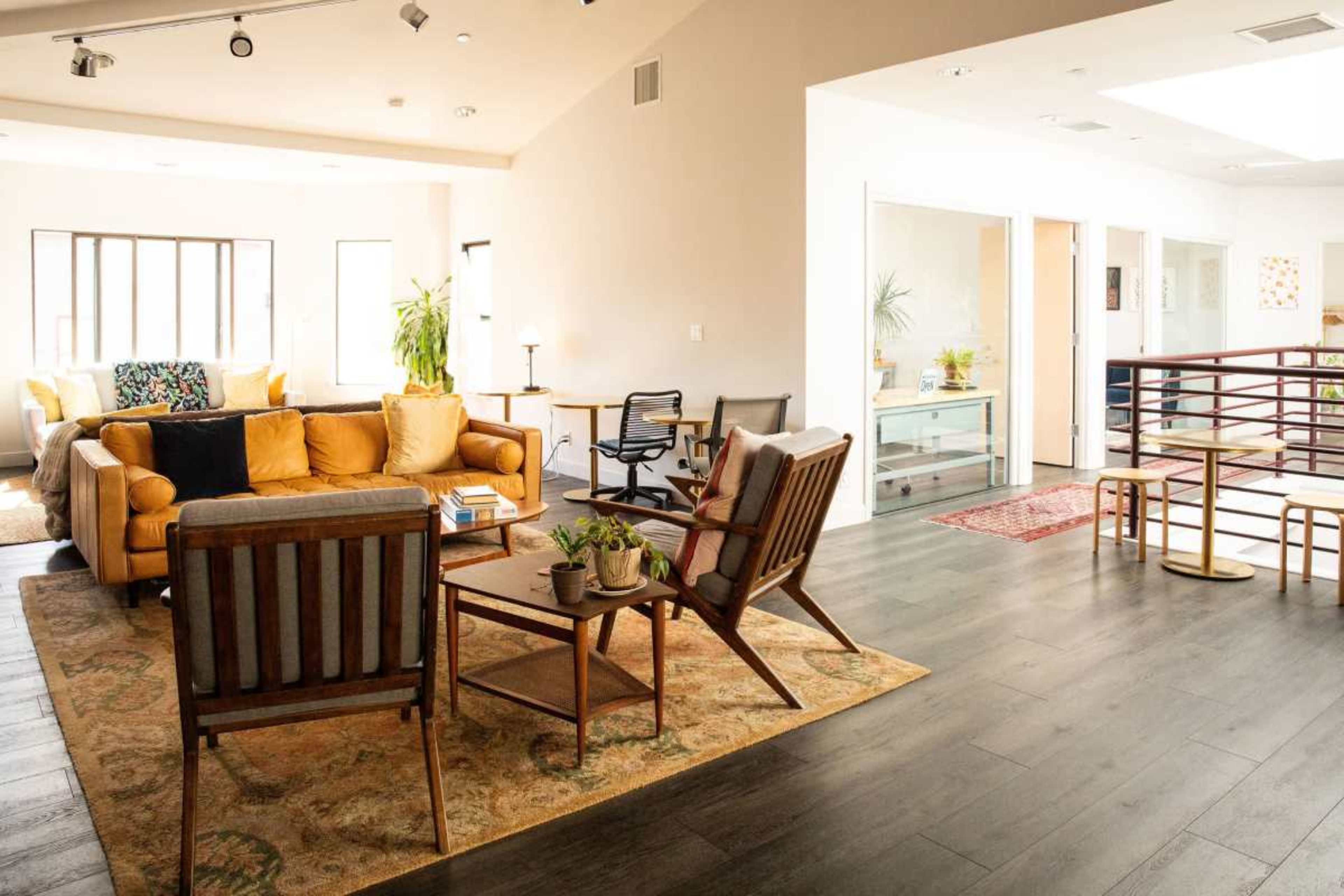 The image shows a spacious, open living area with yellow couches, wooden chairs, a coffee table, and a rug, alongside large windows that let in natural light.