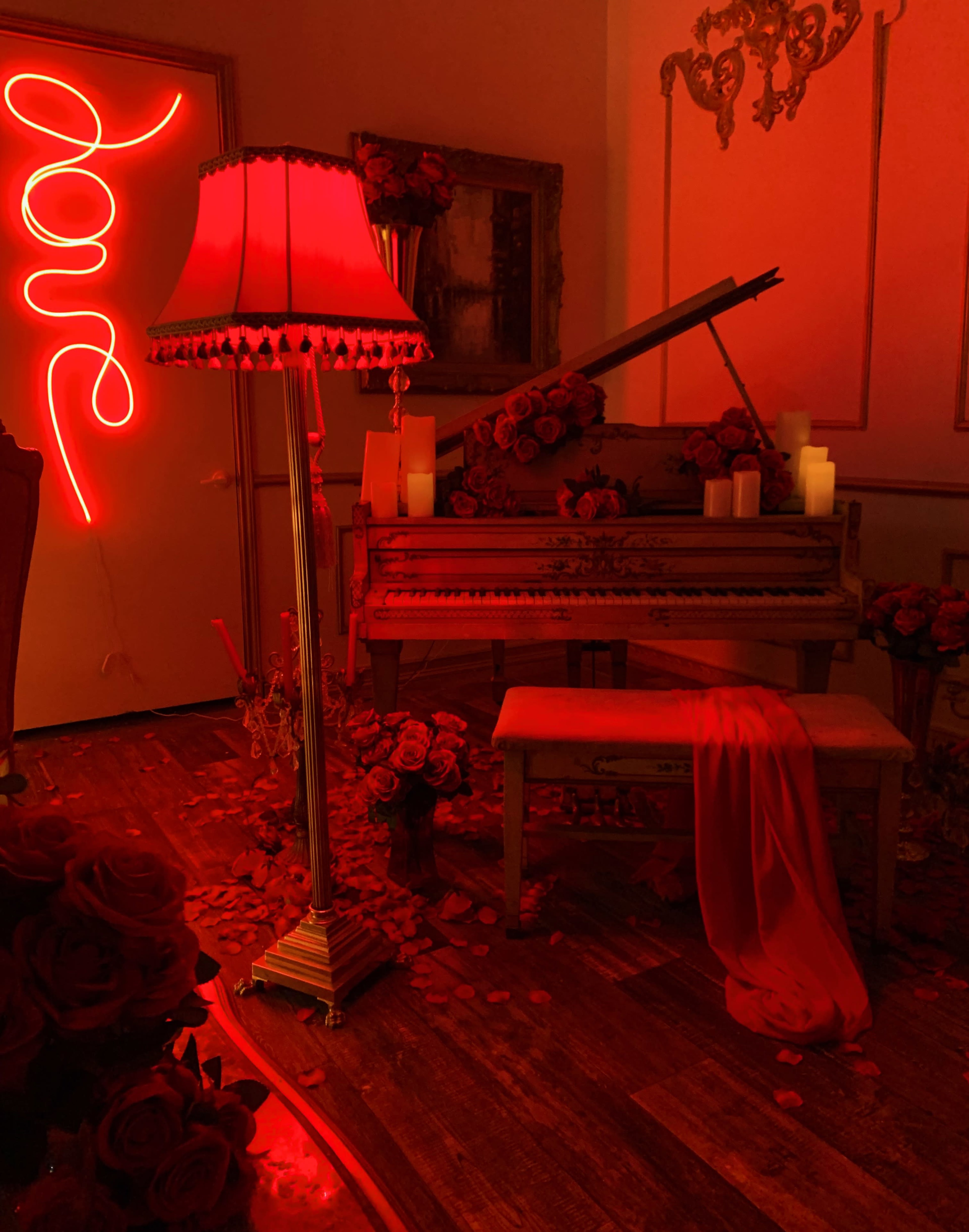 A red-lit room features a vintage piano adorned with roses, candles, and a flowing red fabric.