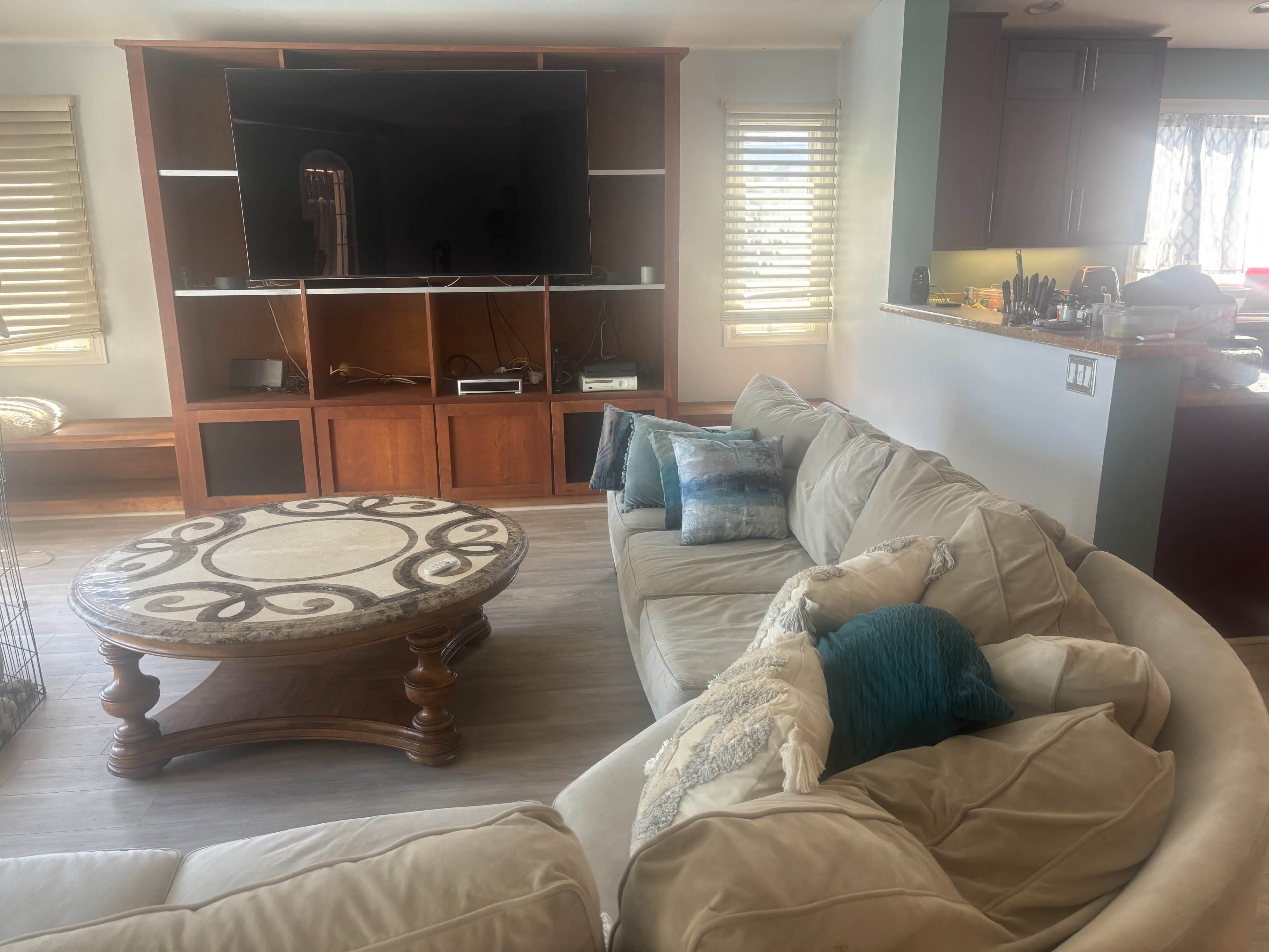 A beige sectional sofa is arranged around a round coffee table in a well-lit living room with a large television mounted on the wall.