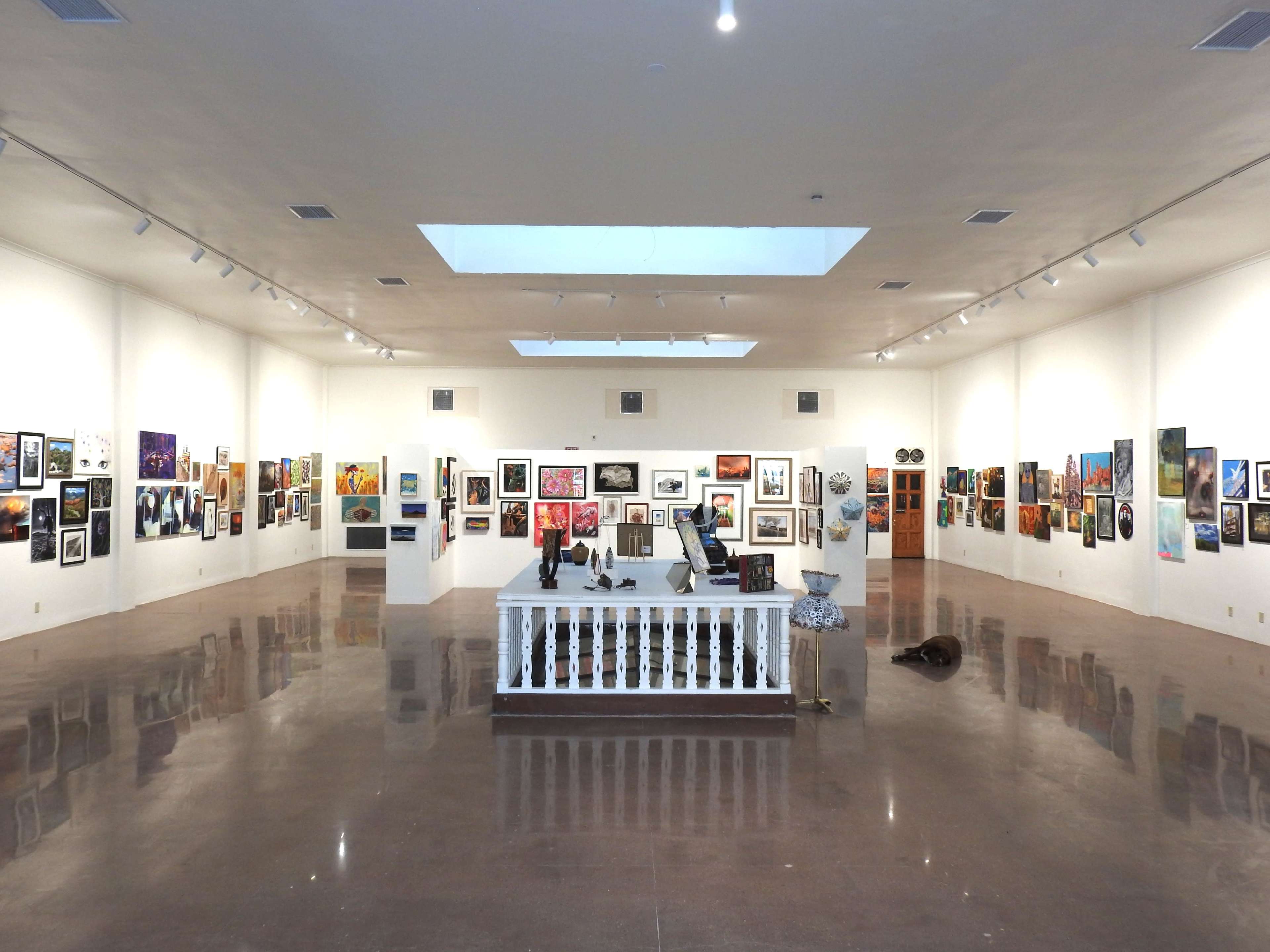 The image shows a spacious art gallery with various artworks displayed on the walls and a central reception area featuring a desk.
