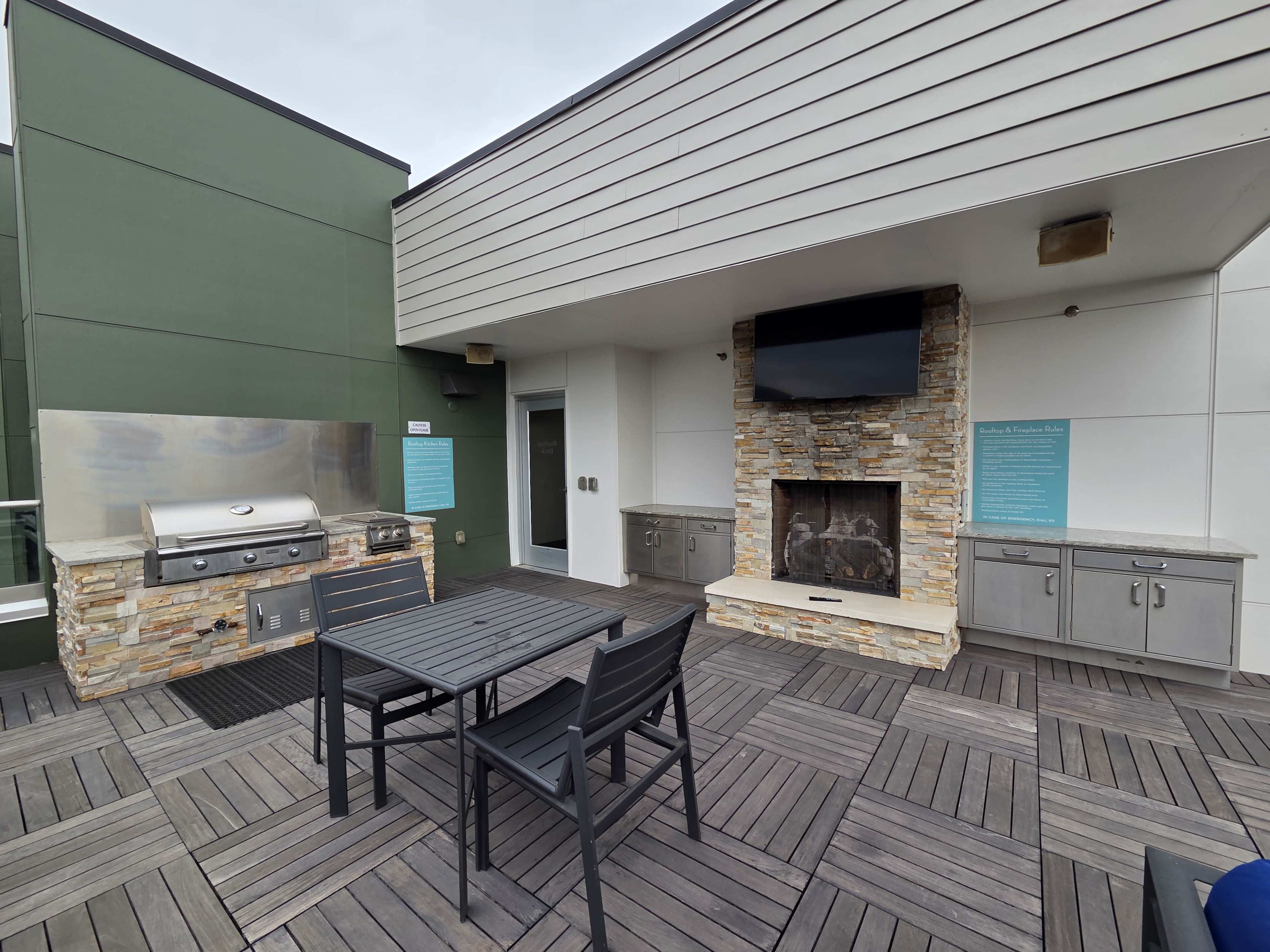 The image shows a rooftop patio featuring a barbecue grill, a stone fireplace, a television, and a dining table with chairs.